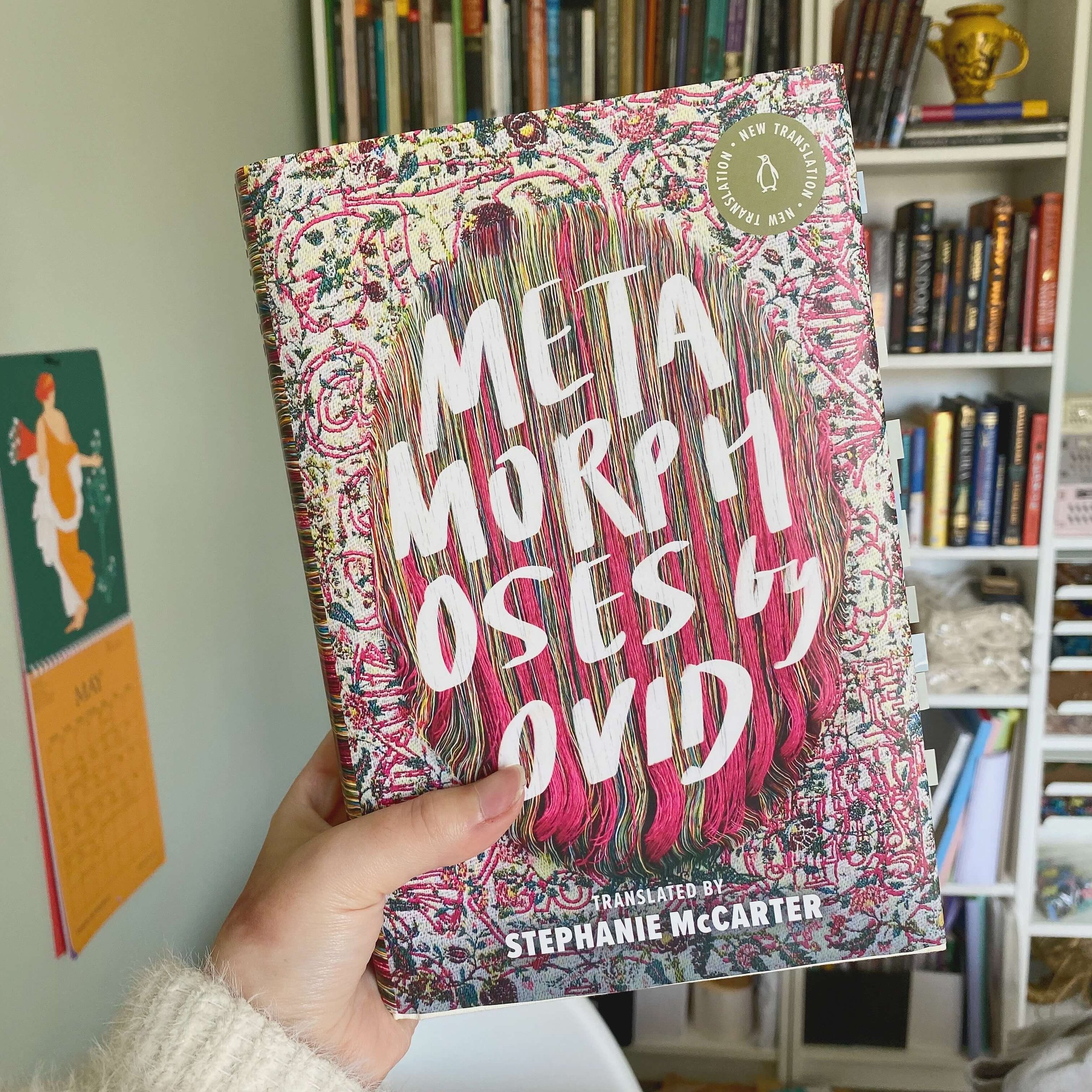 a hand holding up a hardcover book (ovid's metamorphoses, translated by stephanie mccarter) in front of a full bookcase