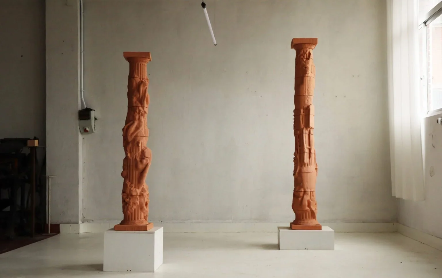 Two tall, intricately carved 3D printed clay terracotta pillars on white pedestals in a minimal room with natural light from a window.