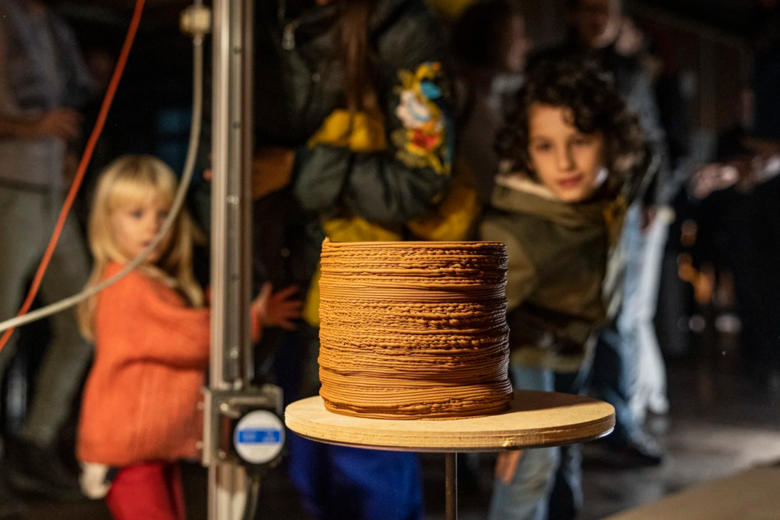 A 3D Printed clay vase with people in the background, including a young girl in an orange sweater and a curly-haired boy looking at the cake.