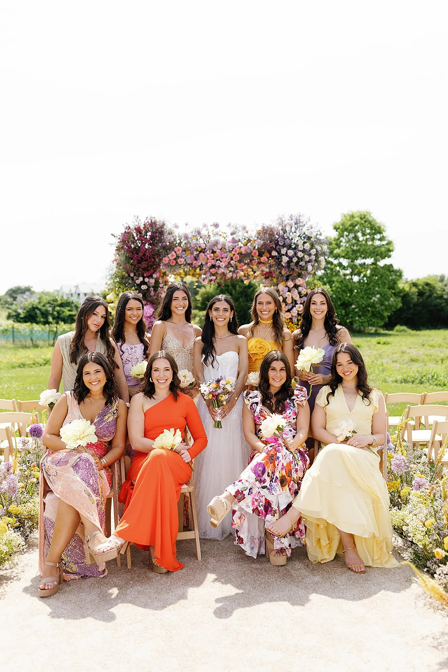  bridesmaids sit around bride in colorful dresses surrounded by vibrant summer florals  