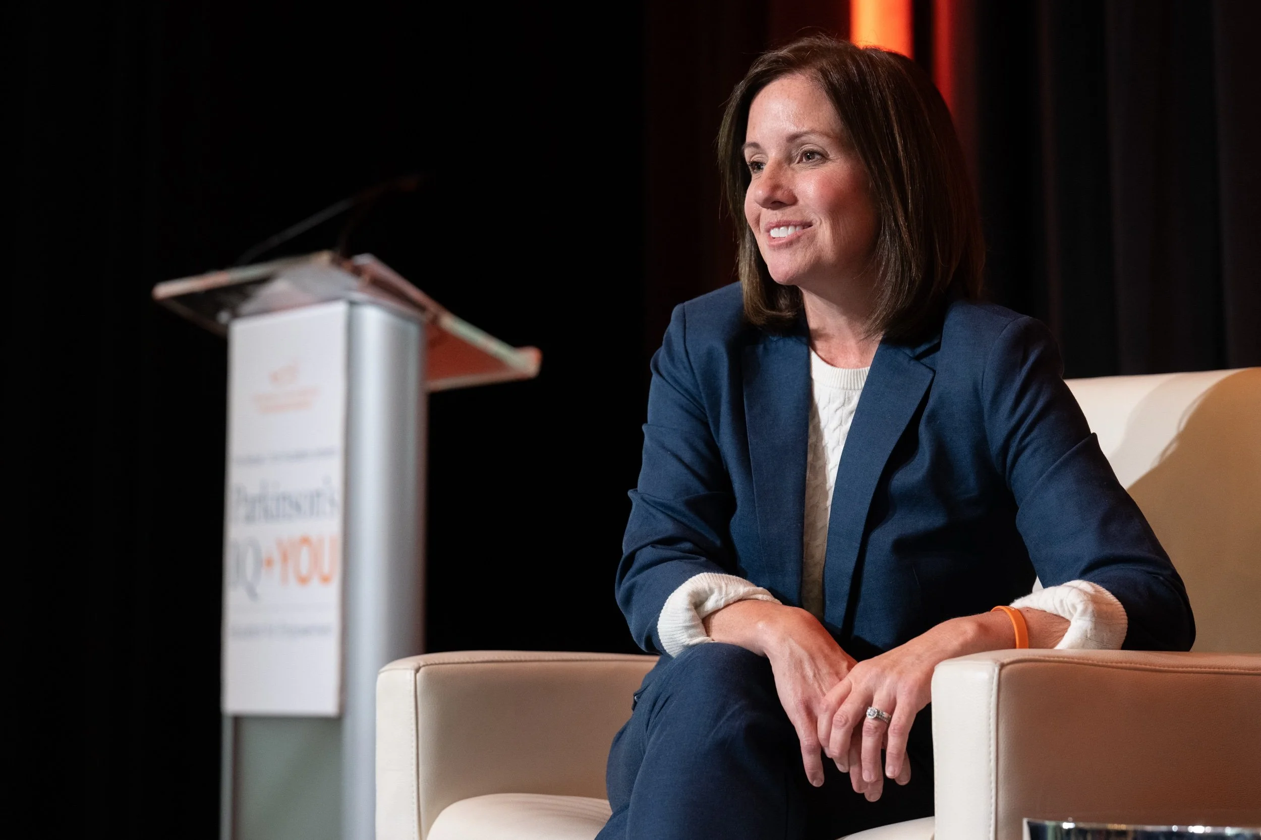 A woman with shoulder-length brown hair, wearing a navy blazer and cream sweater, sitting on a white armchair on a stage, smiling, with a blurred podium to the left.