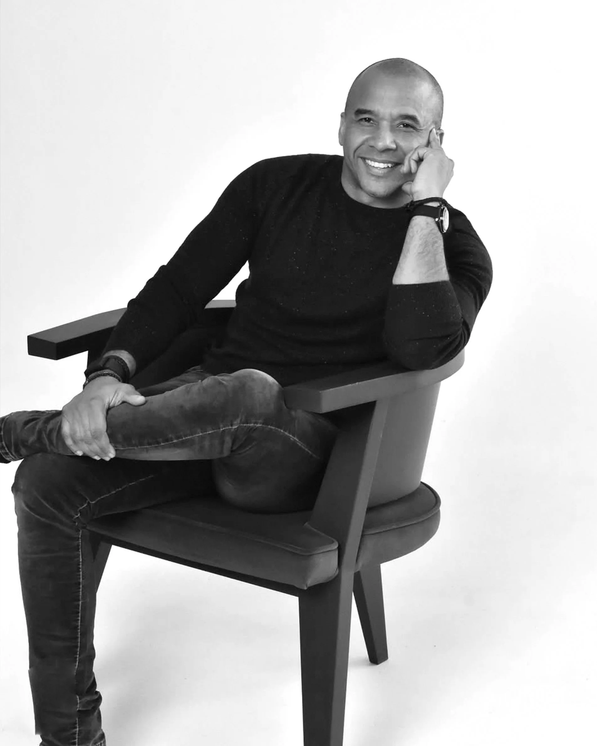 A black and white photo of a smiling man sitting on a wooden chair, resting his head on his left hand, wearing a dark sweater, jeans, and a watch, against a plain white background.