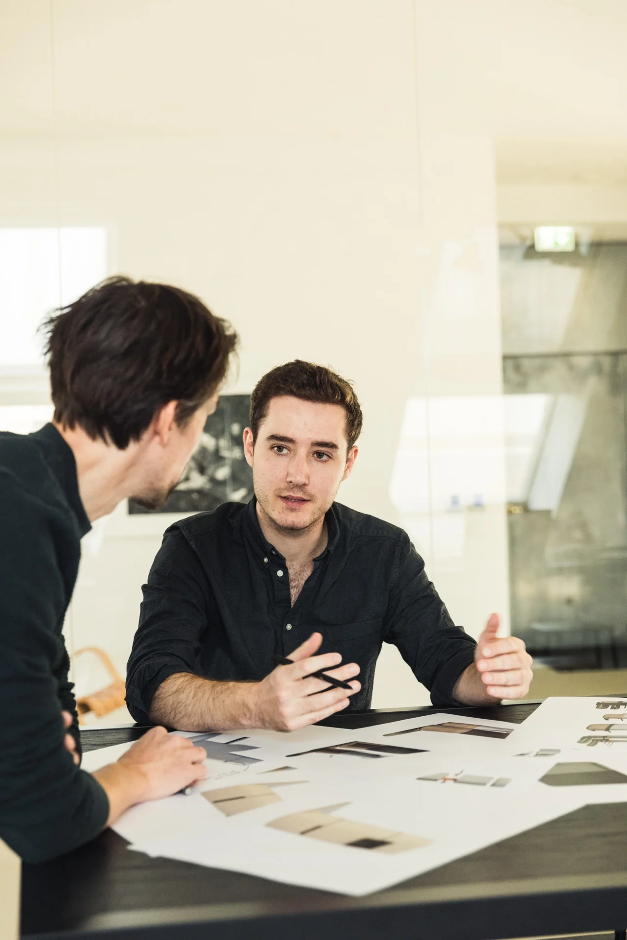 Two men discussing industrial design proposals in a meeting with printed design proposals laid out.