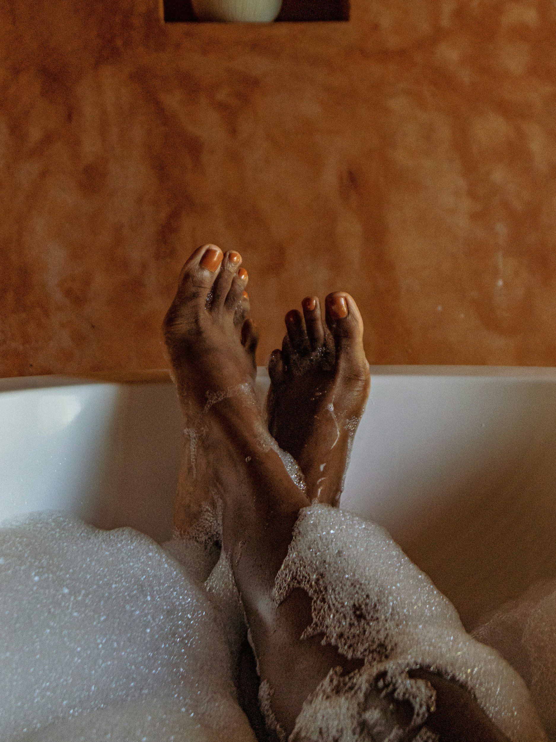 Brown legs and feet extend and rest on the edge of a white bathtub with bubbles floating on the water