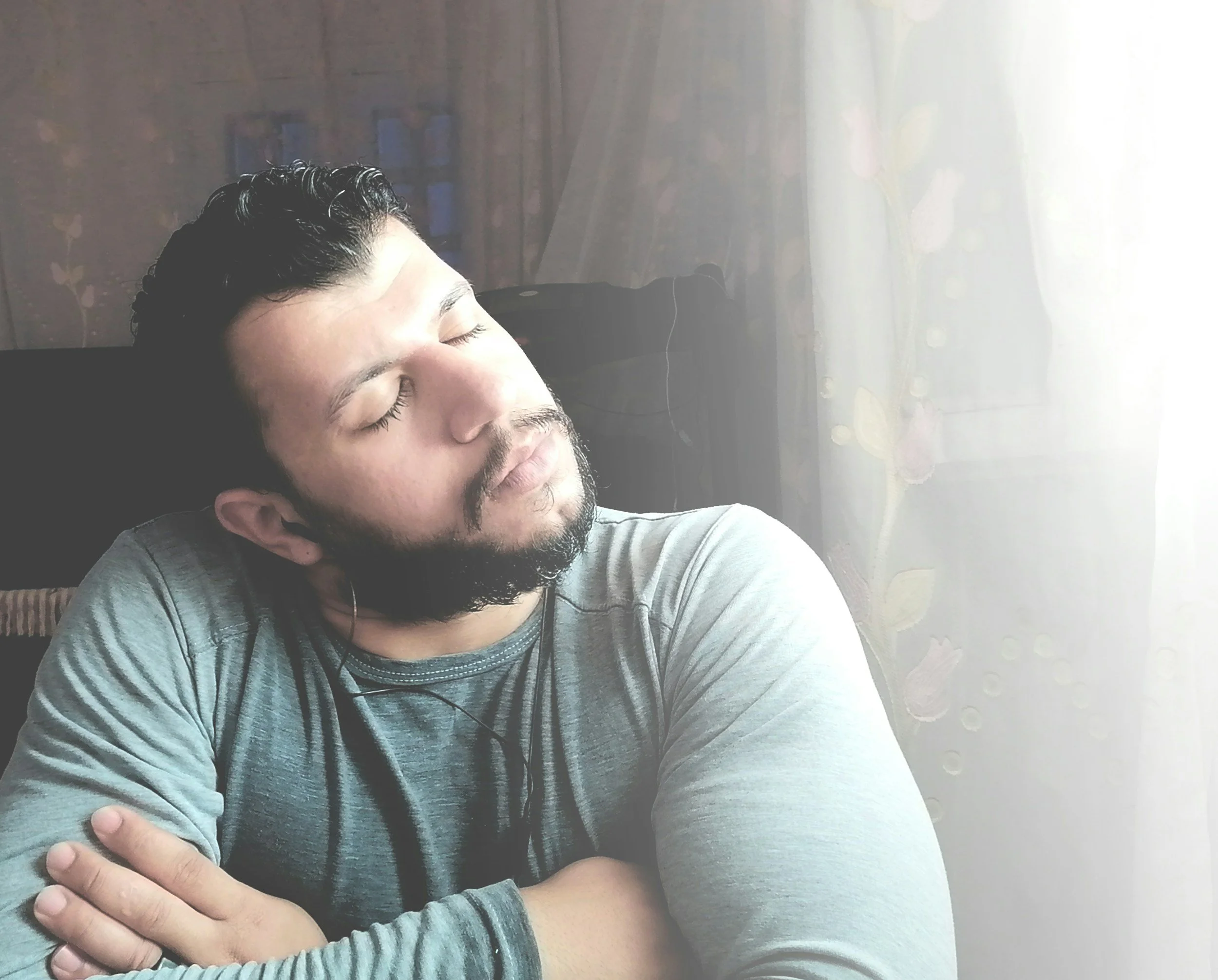 A man sits with arms crossed, looking out a window in quiet reflection, illustrating emotional withdrawal and mental fatigue associated with dad burnout.