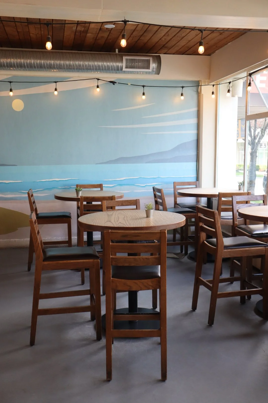 Empty cafe with wooden tables and chairs, small potted plants on tables, painted seaside mural on wall, string lights hanging from ceiling, and large window letting in natural light.