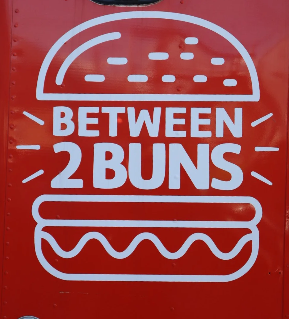 Sign with a stylized hamburger illustration and the text 'Between 2 Buns' in bold letters, indicating a food-related message or brand.