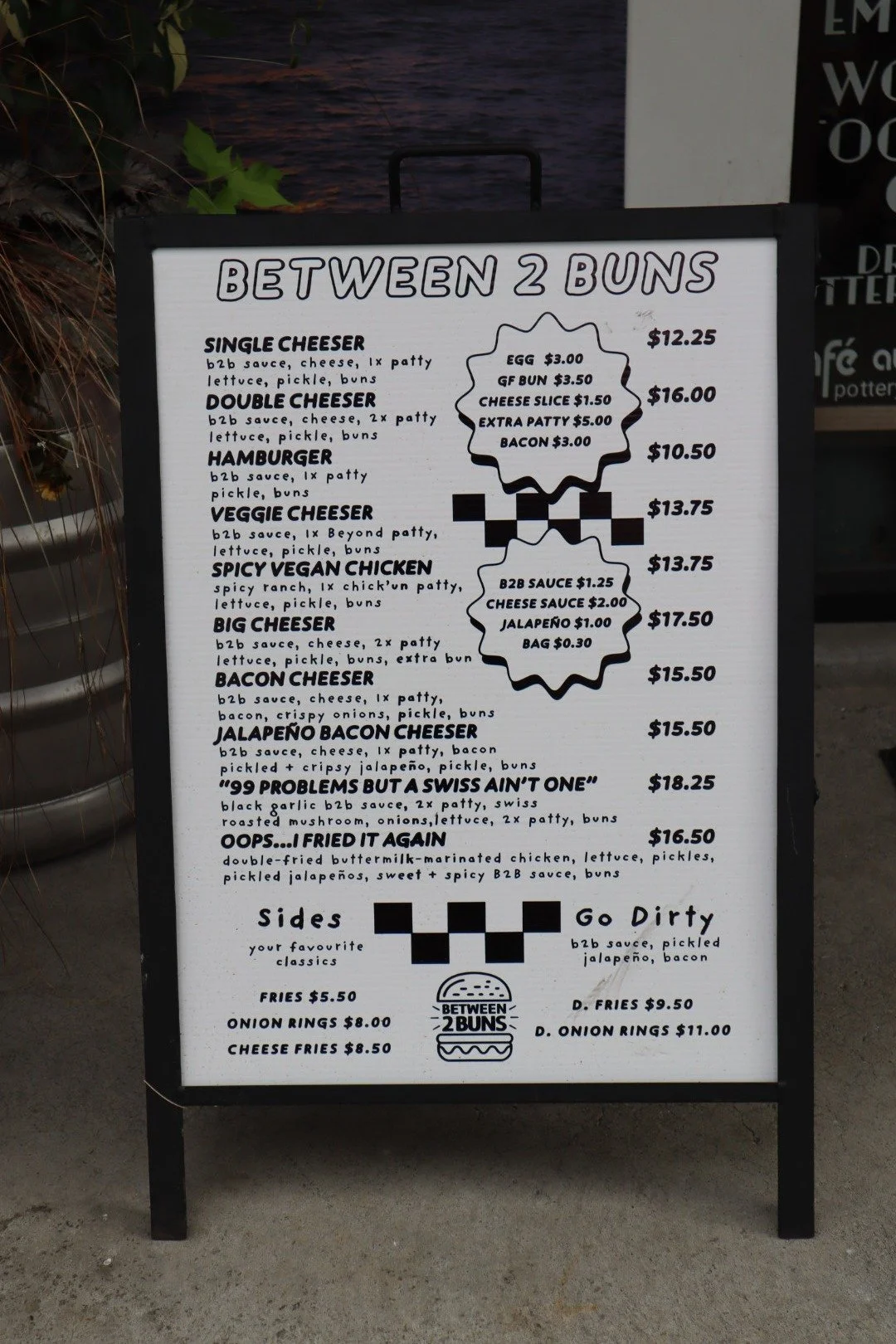 A menu board from 'Between 2 Buns' featuring burgers and sides with prices, including options like single and double cheeser, veggie cheeser, spicy vegan chicken, big cheeser, bacon cheeser, jalapeno bacon cheeser, and specialty burgers with additional charges for extras. Sides include fries, onion rings, and cheese fries, with prices ranging from $5.50 to $11.00.