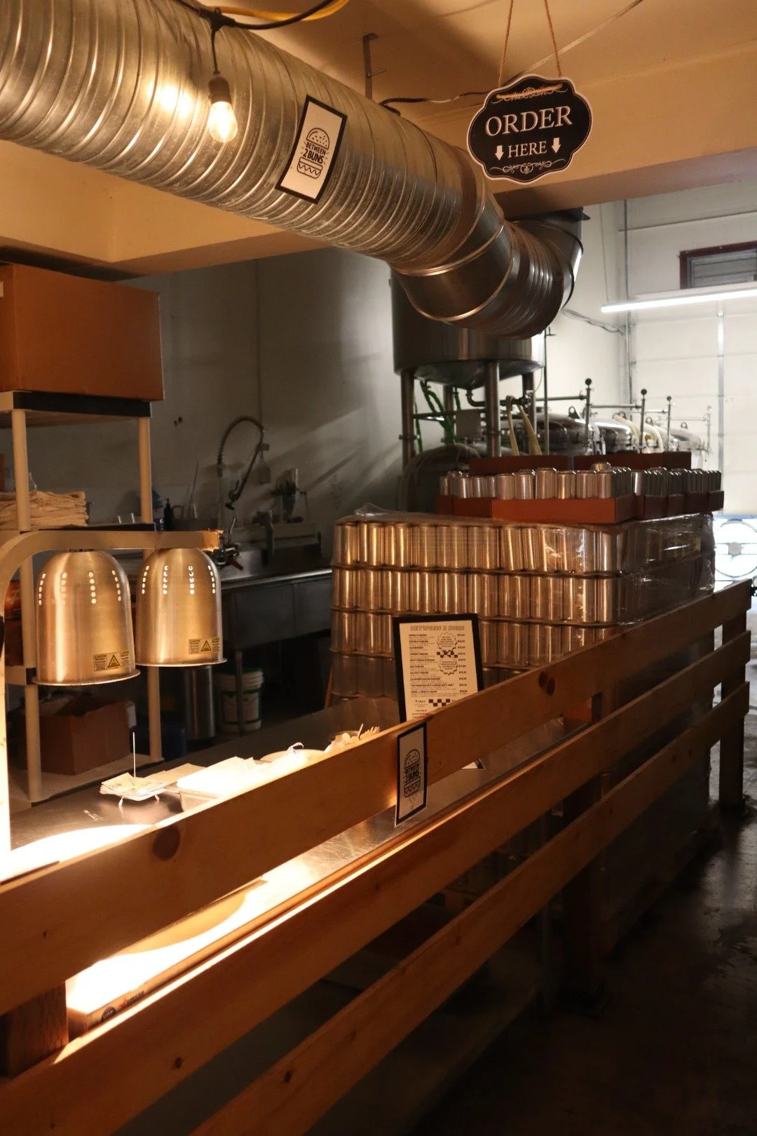 Interior of a coffee shop or cafe with a wooden counter, stacked metal cups, a sign that says 'Order Here,' and industrial piping on the ceiling.