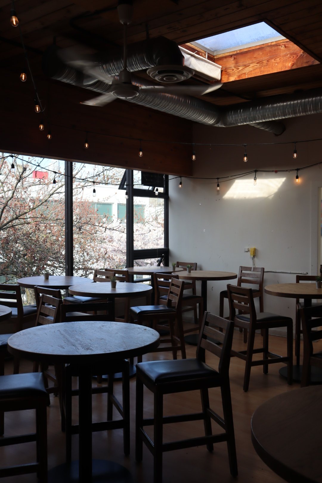 Interior of a cozy cafe with round wooden tables, chairs, string lights hanging from the ceiling, large windows with a view of trees outside, a wooden ceiling with a skylight, and an exposed ventilation duct.