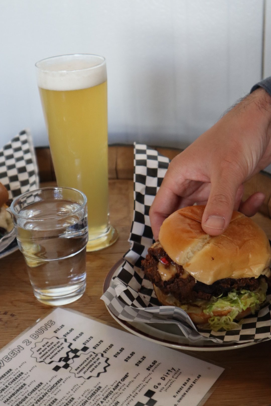 A cheeseburger with lettuce, tomato, and chili, a glass of water, a tall glass of beer, and a menu on a wooden table in a casual restaurant.