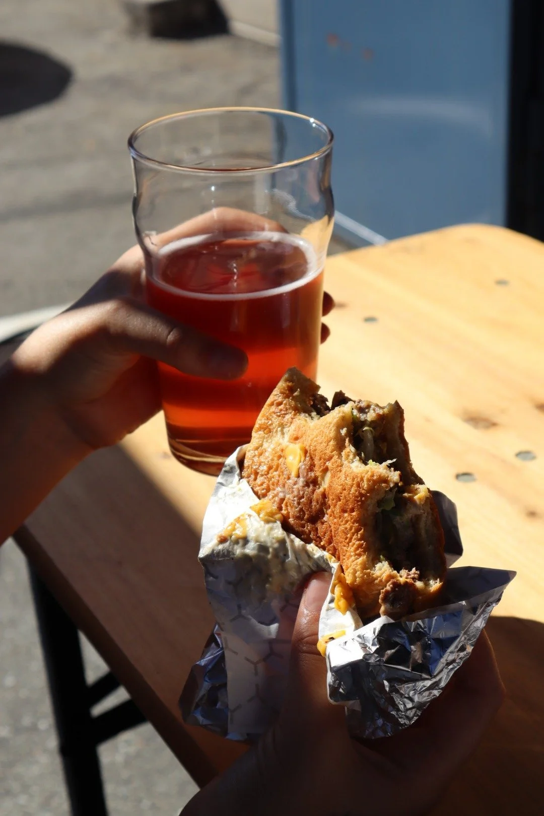 A person holding a glass of iced tea in one hand and a half-eaten cookie and bun in the other, with a wooden table and outdoor background.