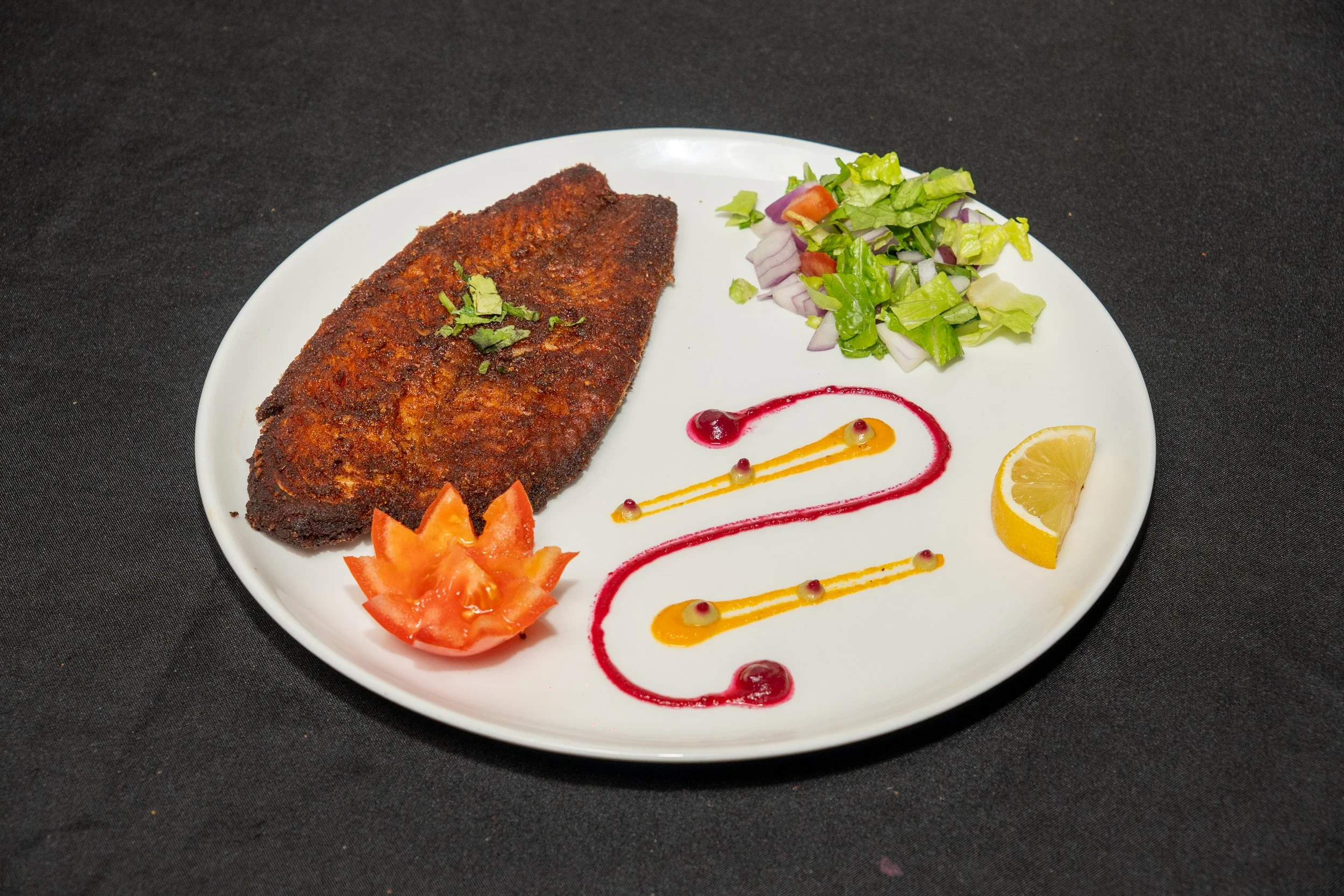Plate with fried fish, chopped tomato, salad, lemon wedge, and decorative sauces on a black surface.