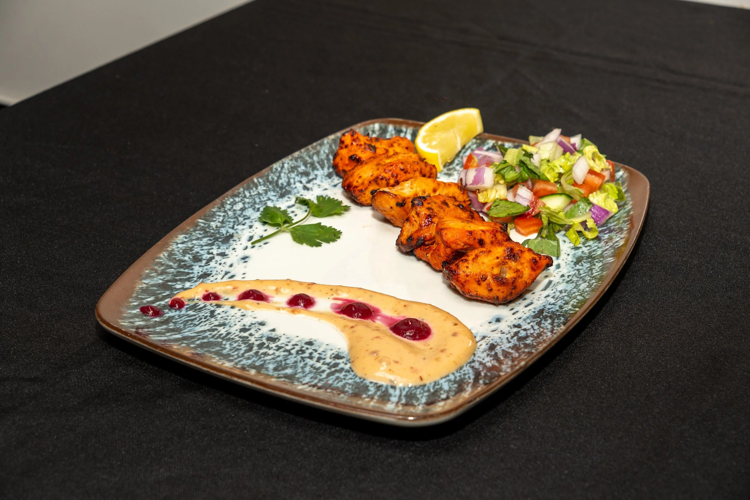 Plate with grilled chicken curry, salad, lemon wedge, and cream with red sauce.