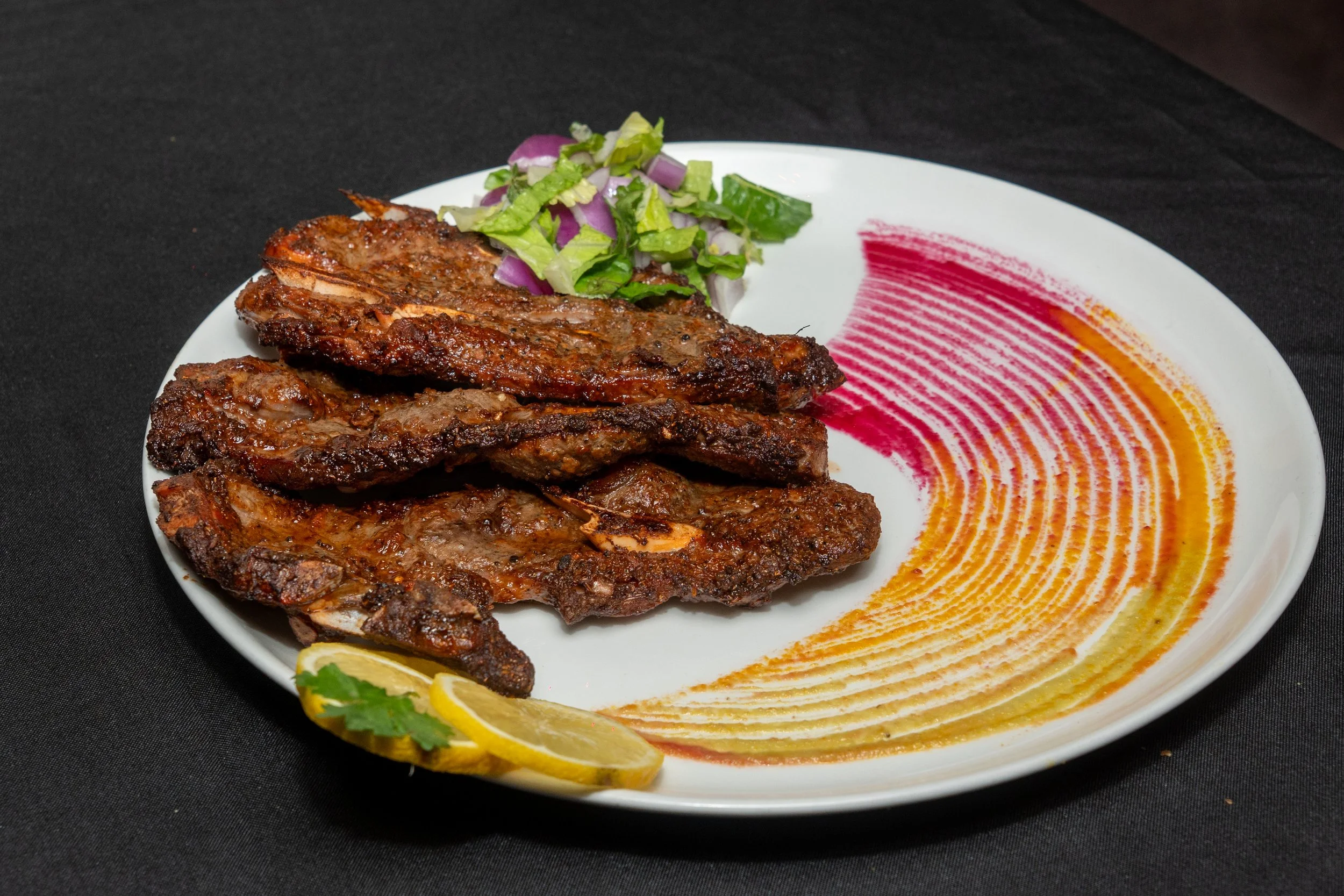 Plate of grilled marinated meat with lemon slices, salad, and colorful sauce streaks