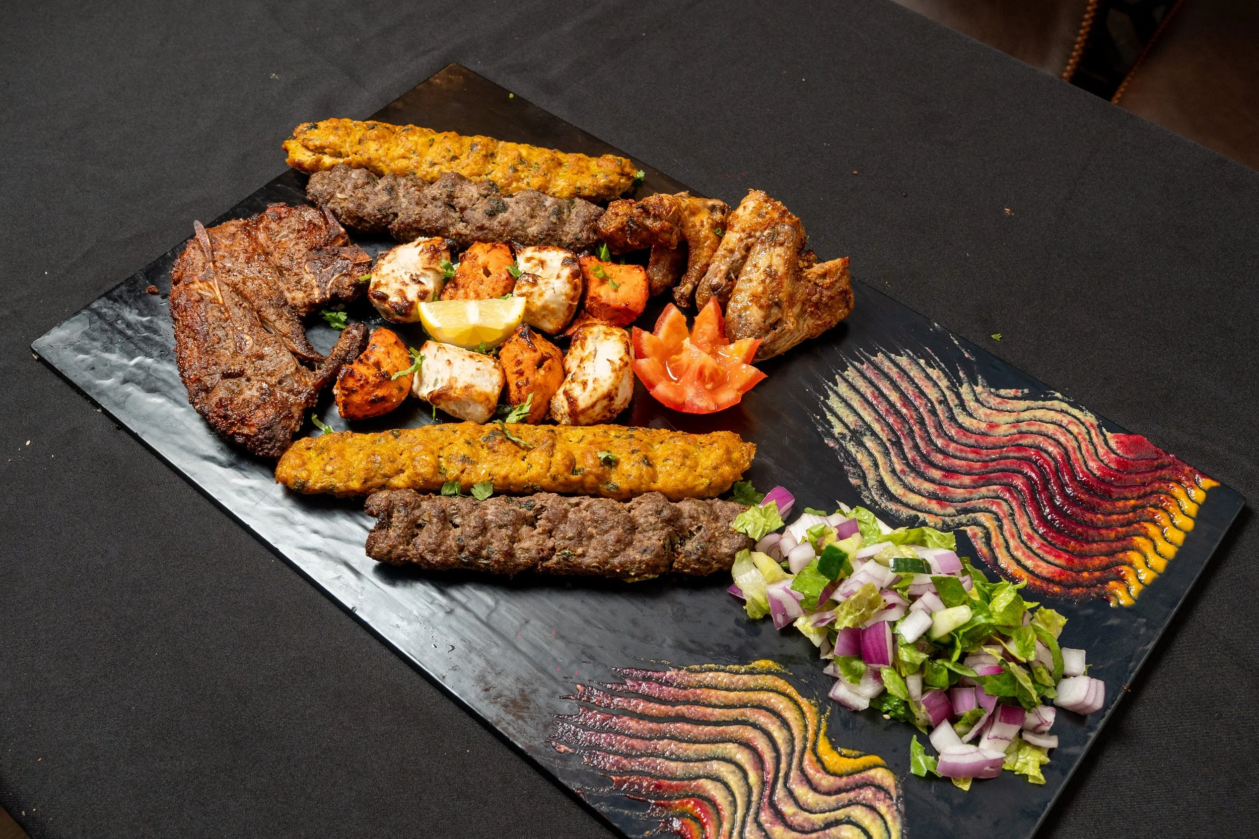 Platter of assorted grilled meats including beef, chicken, and lamb, accompanied by roasted vegetables, chopped onions with cilantro, lemon wedge, sliced tomatoes, and colorful wavy sauces on a black plate.