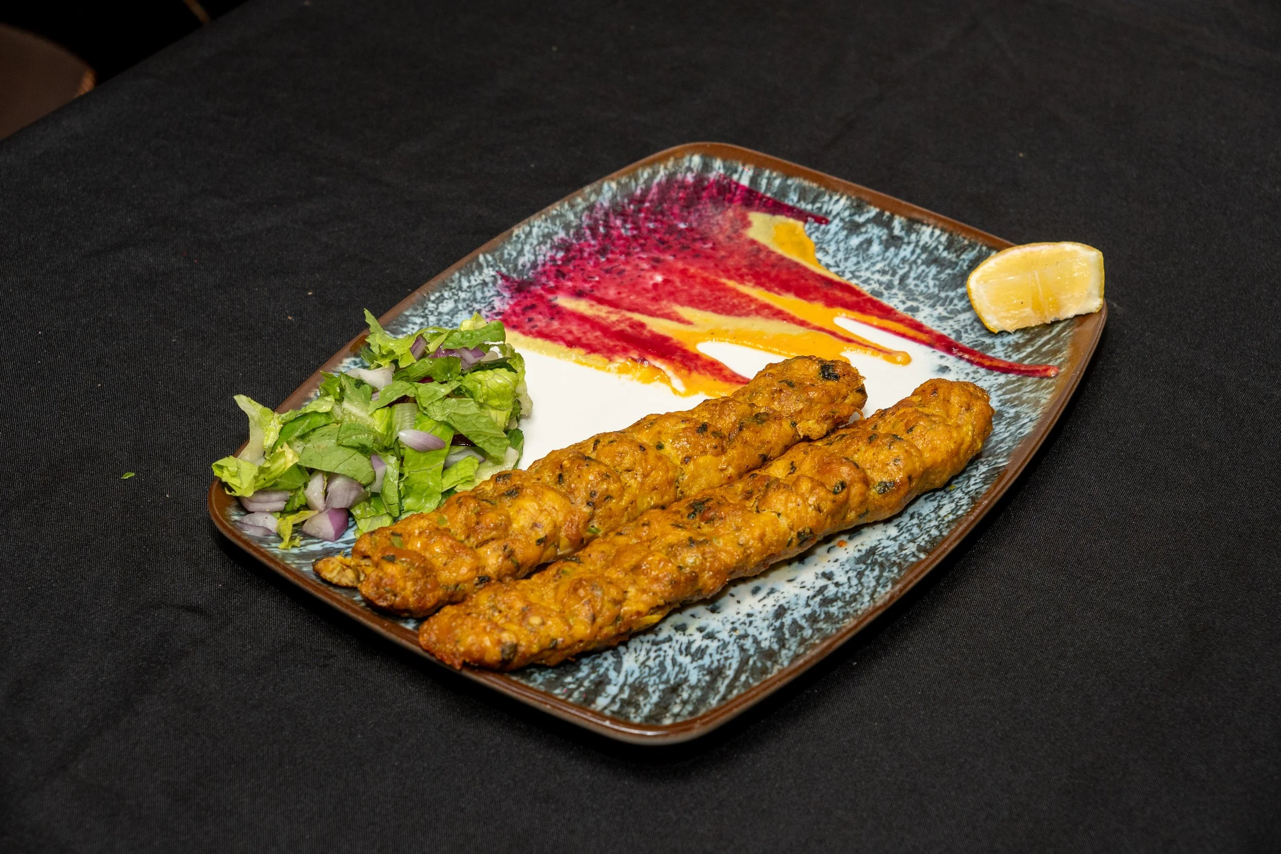 Plate with two long, golden-brown, battered fish fillets, a small mixed green salad with chopped red onions, a slice of lemon, and colorful sauce smear on a decorative rectangular plate placed on a black table.