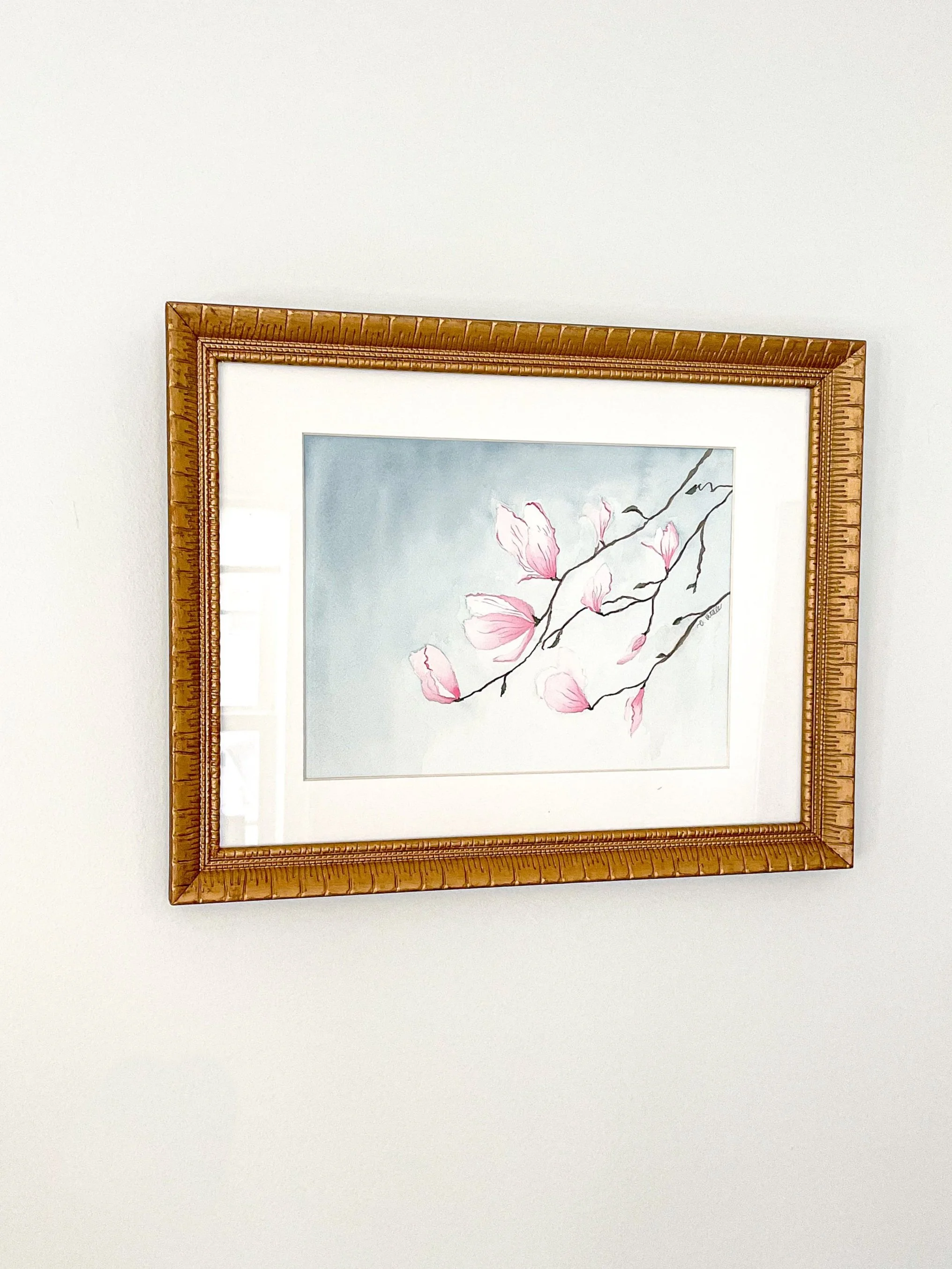 Framed watercolor painting of pink magnolia blossoms on a branch against a light blue background.