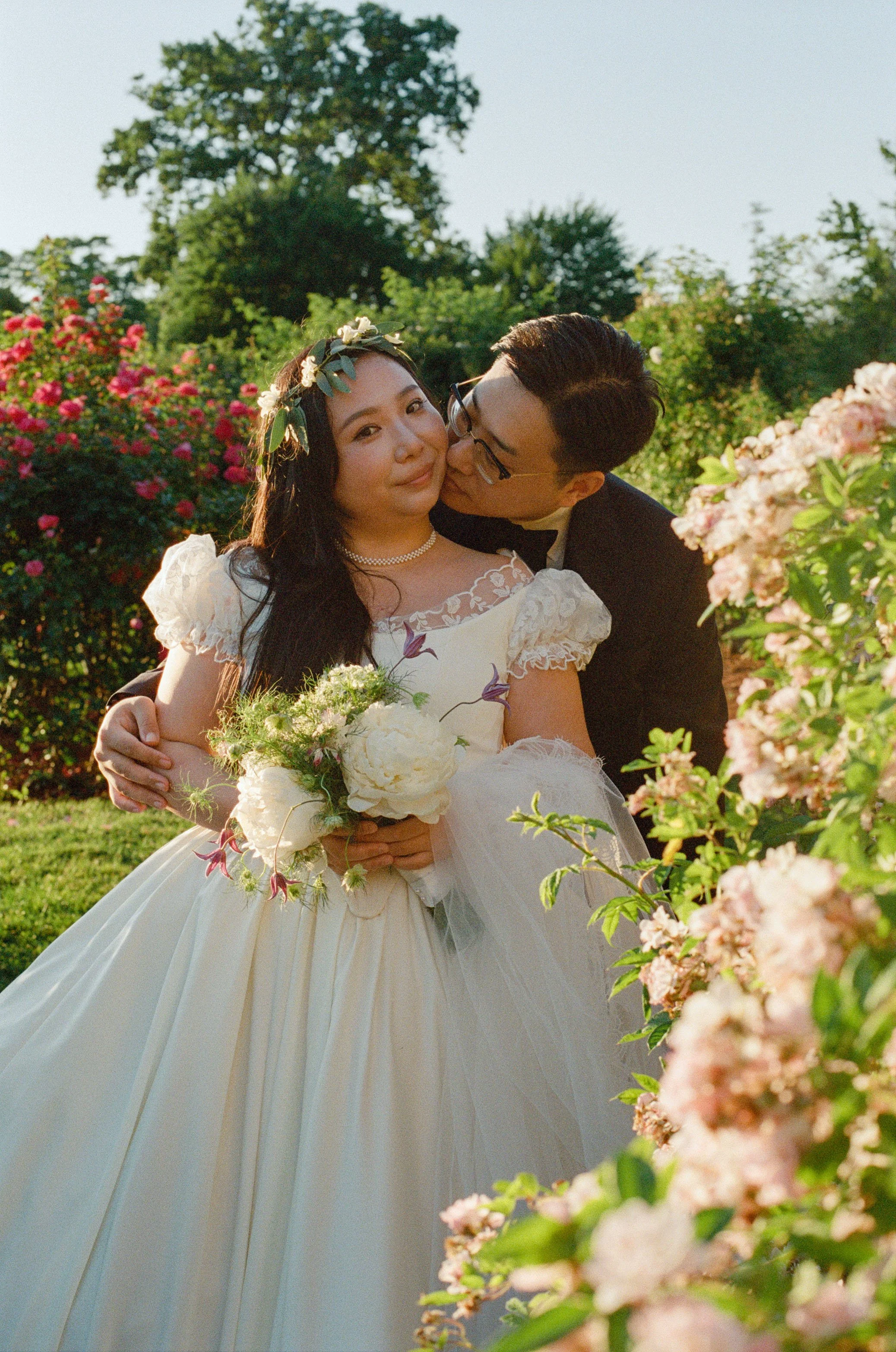 Rose garden wedding on 35mm film. Asian husband kissing his wife on the cheek at Elizabeth Park Rose Garden by Briana Raucci, wedding photographer in Connecticut. Destination wedding on film. Inclusive wedding