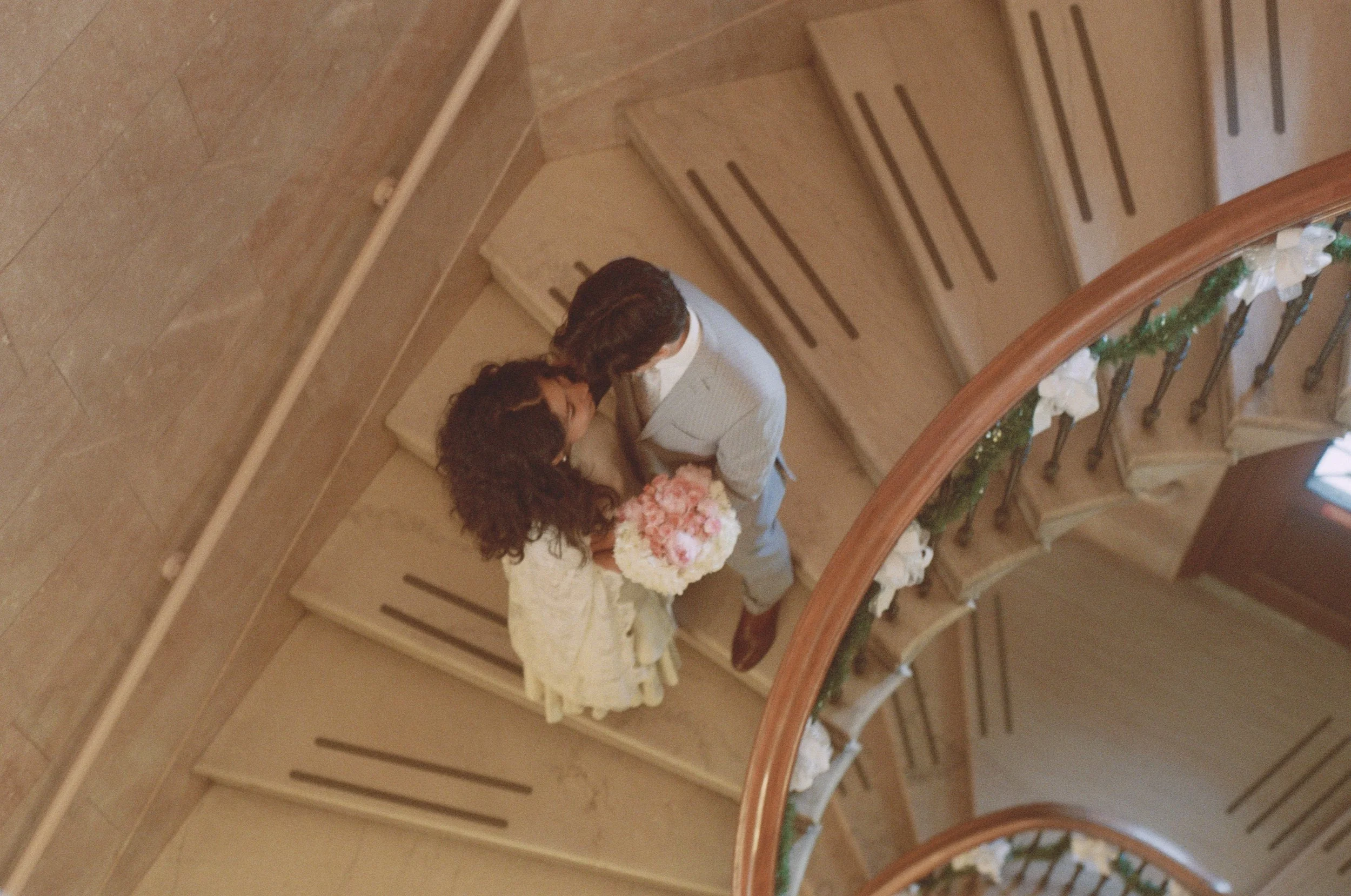 Couple stands on spiral staircase at James Blackstone Library in Branford, CT. Taken by CT film wedding photographer Briana Raucci. Wife is holding a floral arrangement.