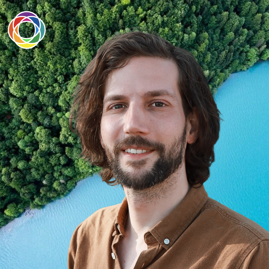 A man with shoulder-length brown hair and a beard smiling in front of a background of dense green forest and a blue body of water.
