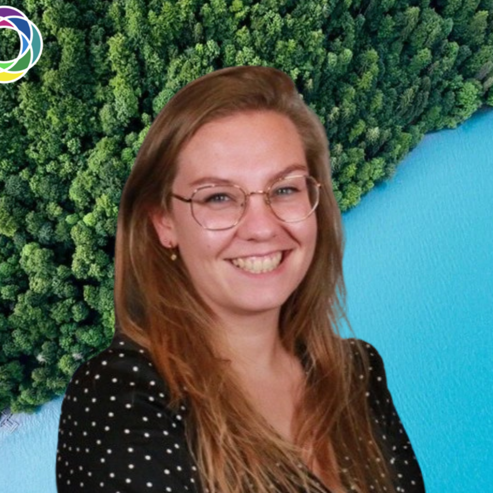 A woman with glasses and long hair smiling in front of a background showing a dense green forest and blue water.