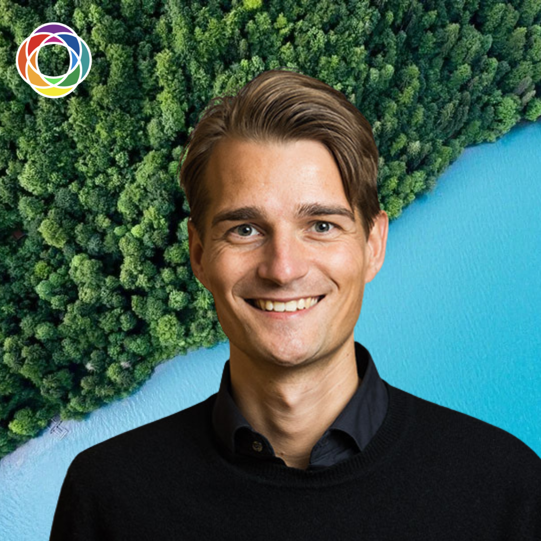 A smiling man with brown hair and blue eyes in front of a background of dense green forest and a blue body of water, with a colorful circular logo in the top left corner.