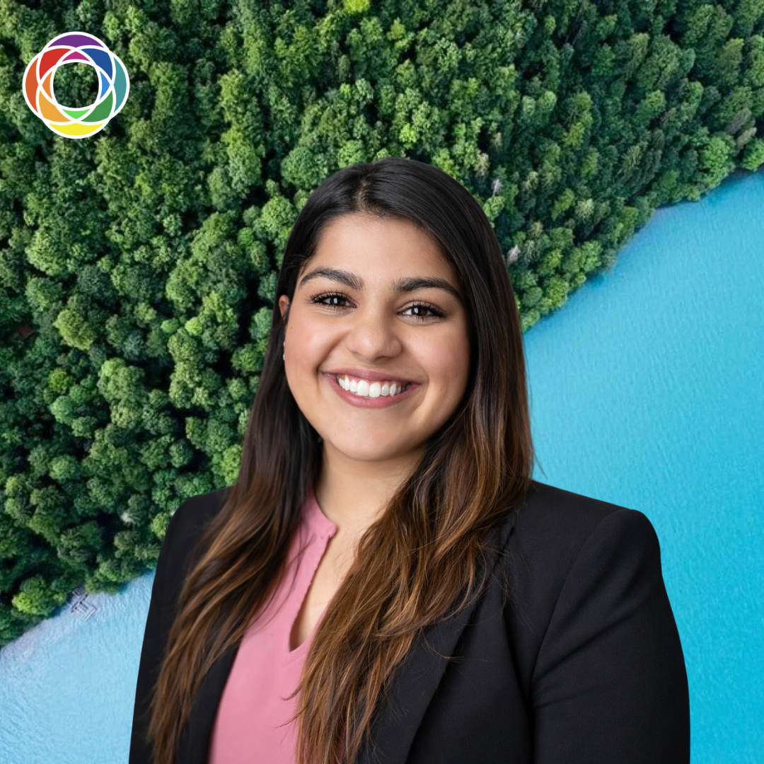A smiling young woman with long brown hair wearing a black blazer and pink top, standing in front of an aerial view of a lush green forest and a blue lake.