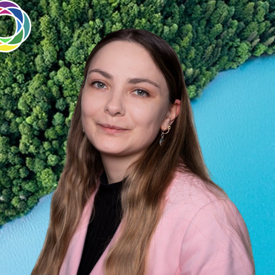 A young woman with long light brown hair, blue eyes, wearing earrings, a black top and pink blazer, standing in front of a background of dense green trees and a blue body of water.