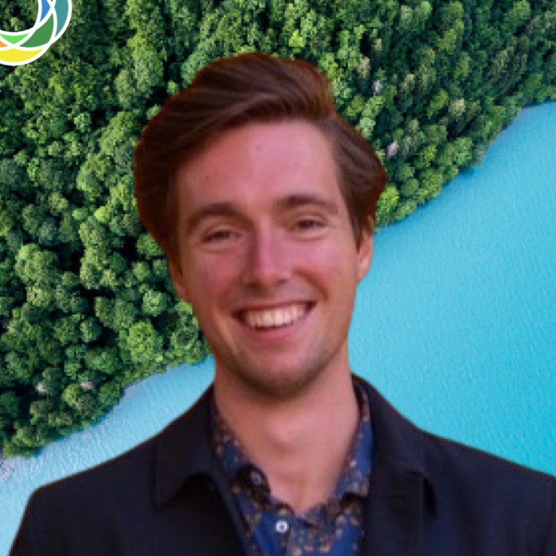 A young man with brown hair and a bright smile, wearing a dark blazer over a patterned shirt, standing in front of a background showing a dense green forest, a light blue body of water, and a partial logo in the top left corner.