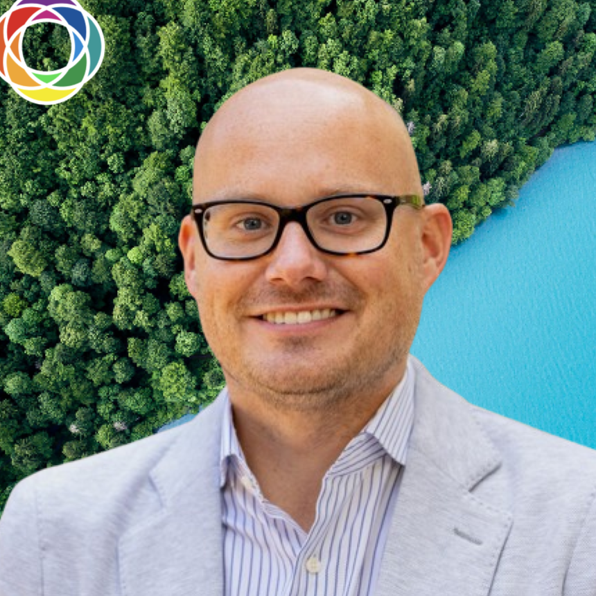 A smiling man with glasses and a striped shirt wearing a light-colored blazer, standing in front of a background featuring a dense green forest and a blue lake.