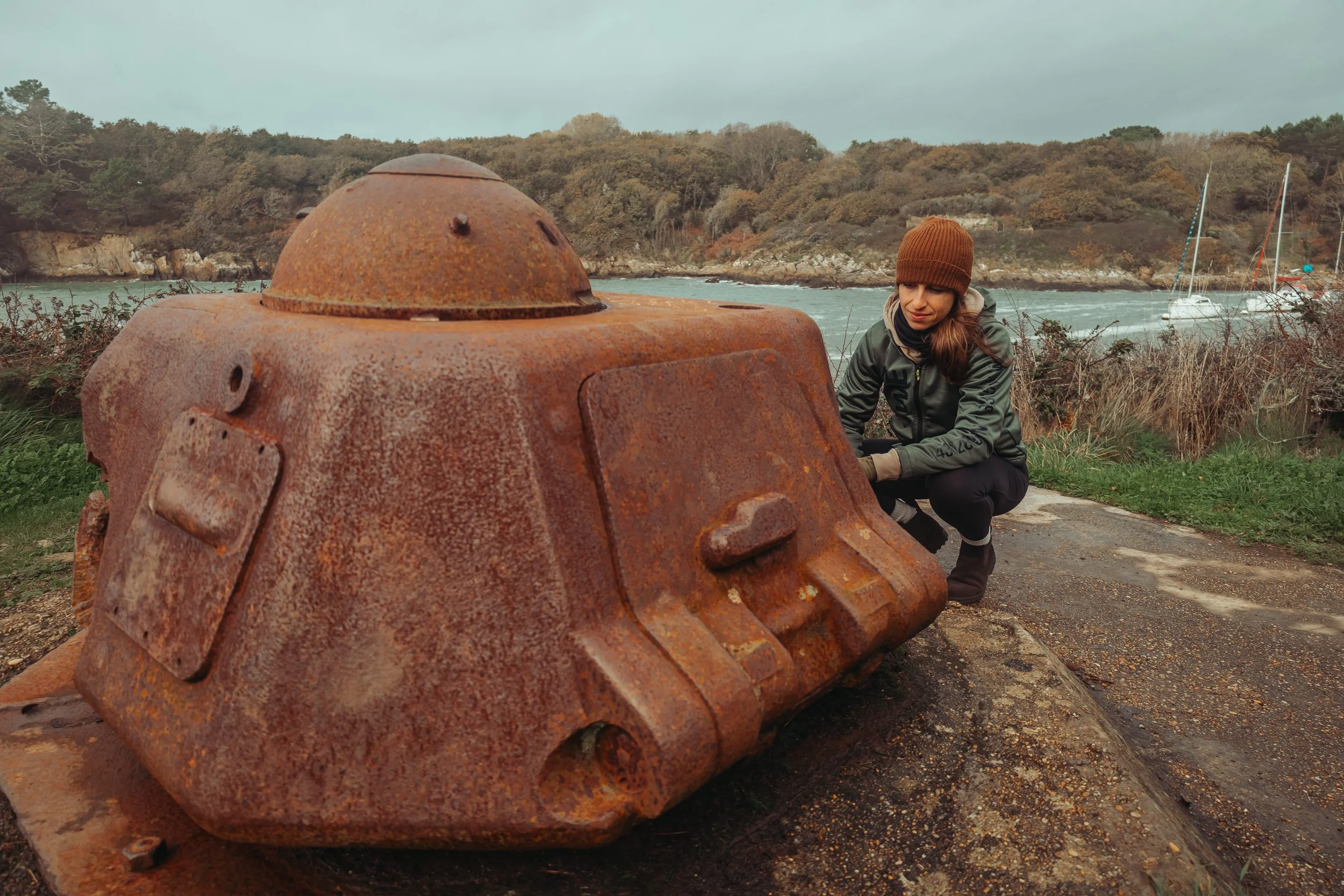 Vue d'une plage rocheuse sous un ciel nuageux, avec une vieille structure en béton ou en métal en premier plan et des maisons modernes en arrière-plan.