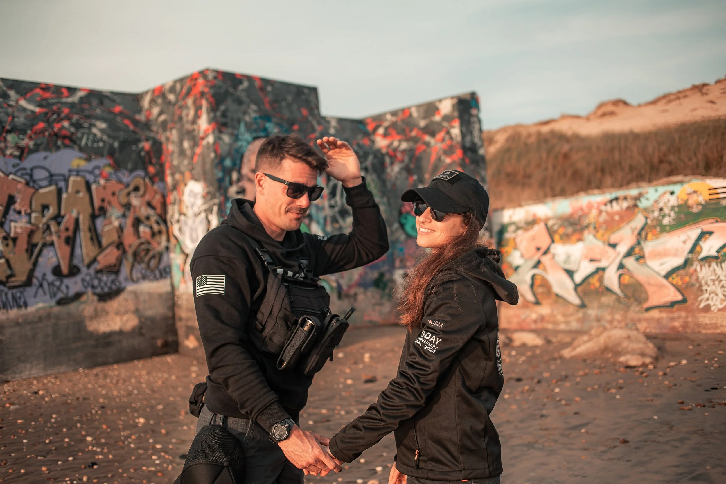 Un couple souriant, tenant la main, debout devant un mur de graffiti sur une plage au coucher du soleil.