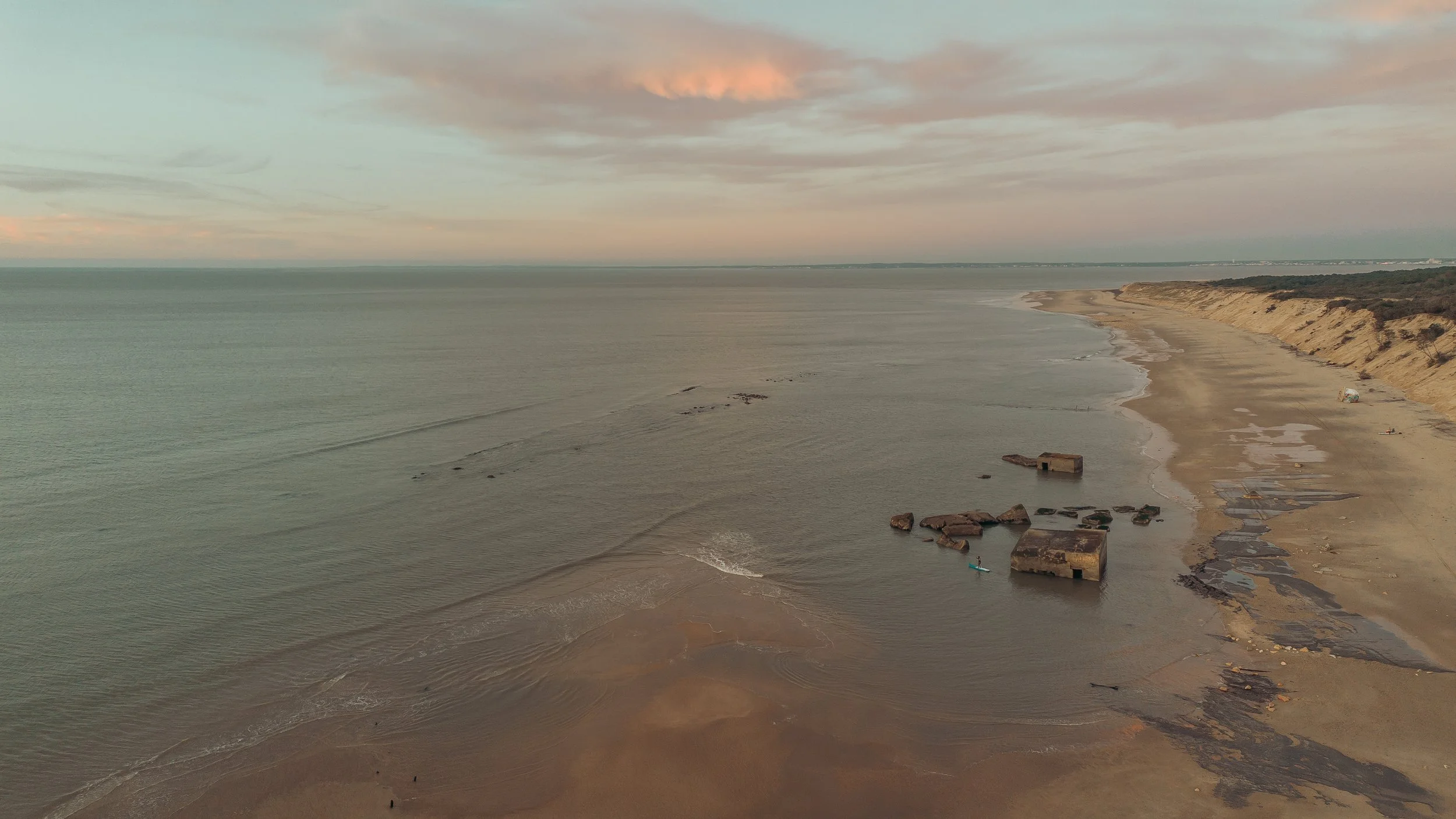 Personnage faisant du paddle devant des rochers à la mer, plage et côte en arrière-plan sous un ciel clair.