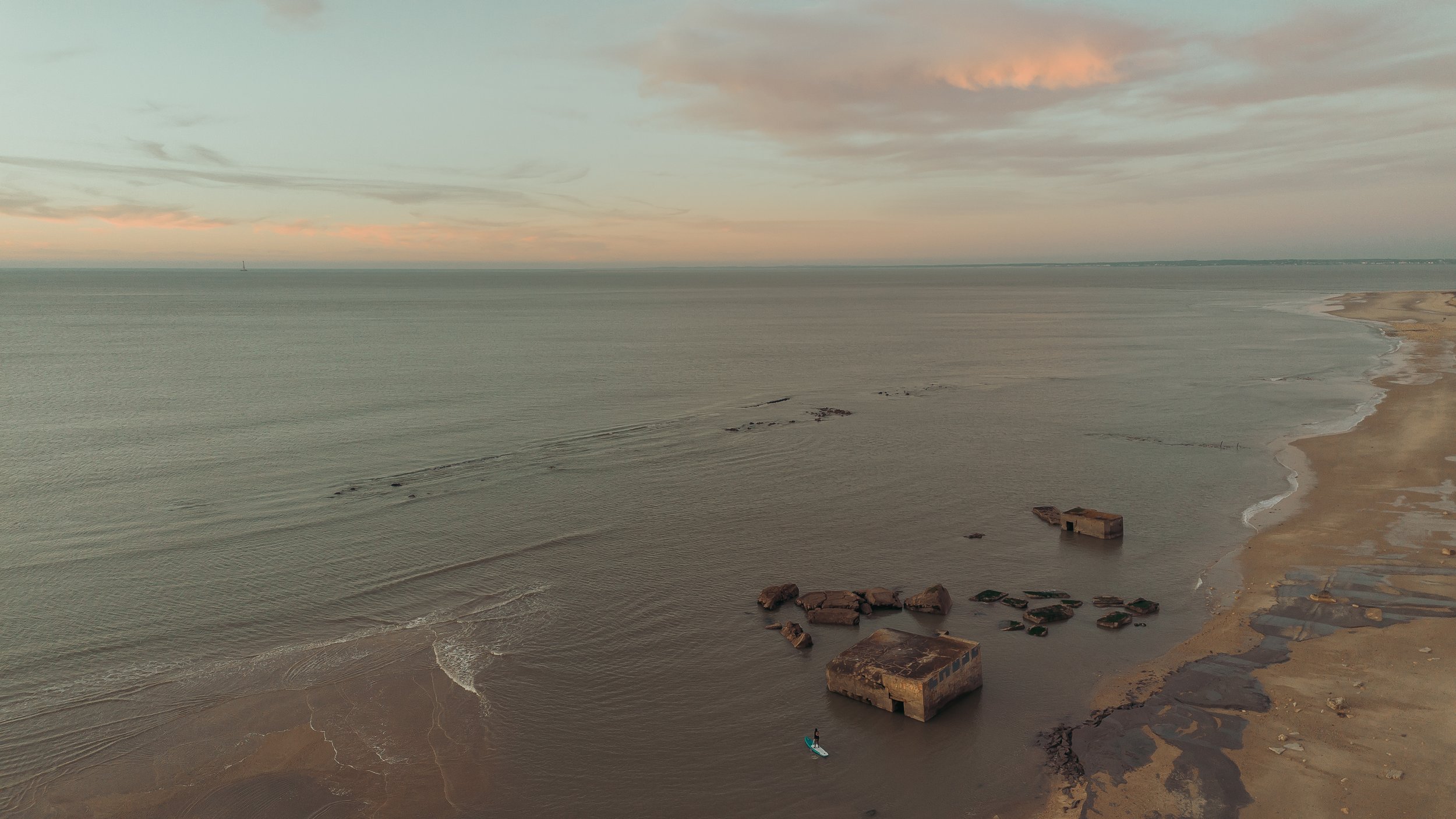 Personnage faisant du paddle devant des rochers à la mer, plage et côte en arrière-plan sous un ciel clair.