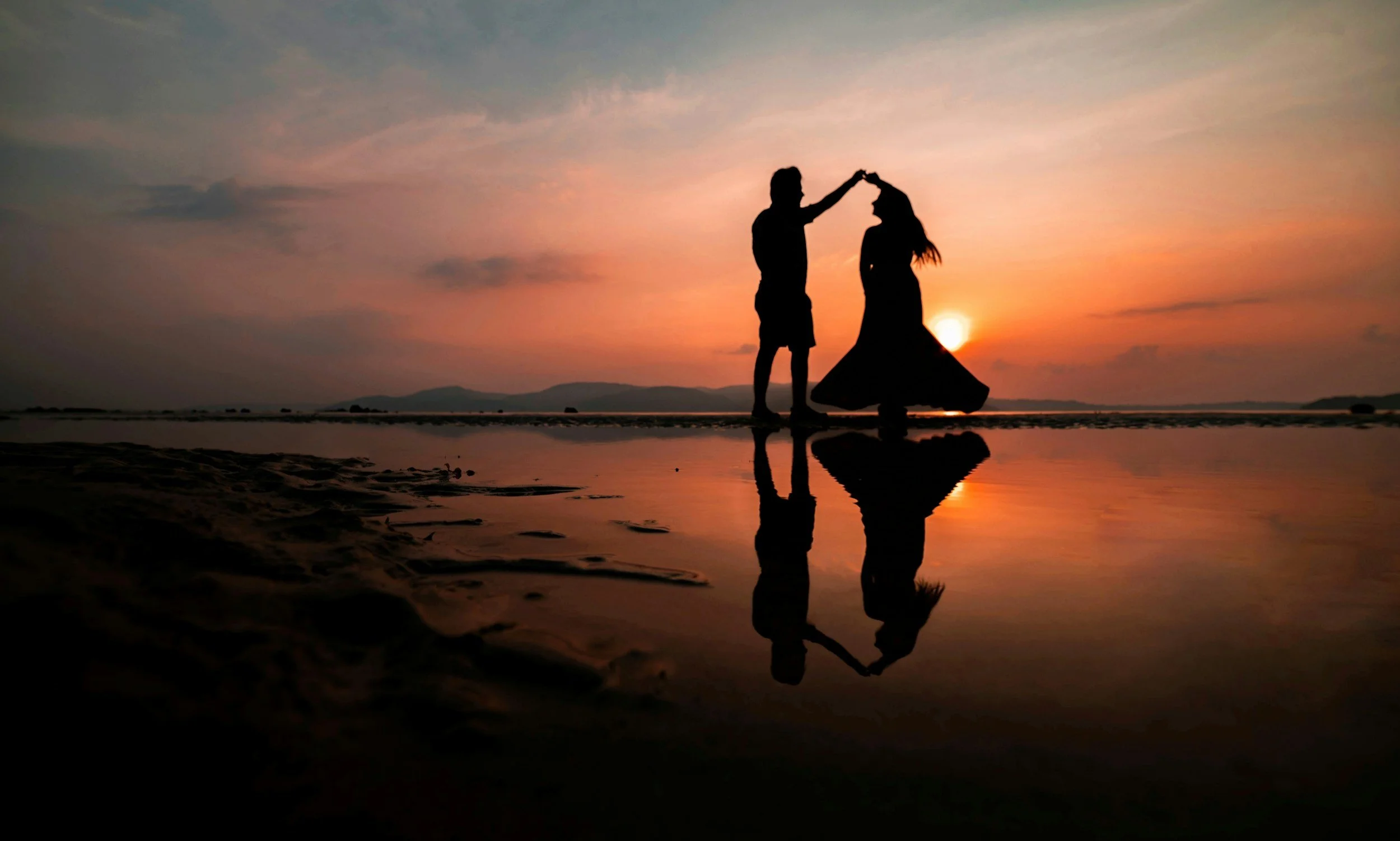 Silhouettes of a man and woman dancing on a beach during sunset, with their reflections visible on the wet sand.