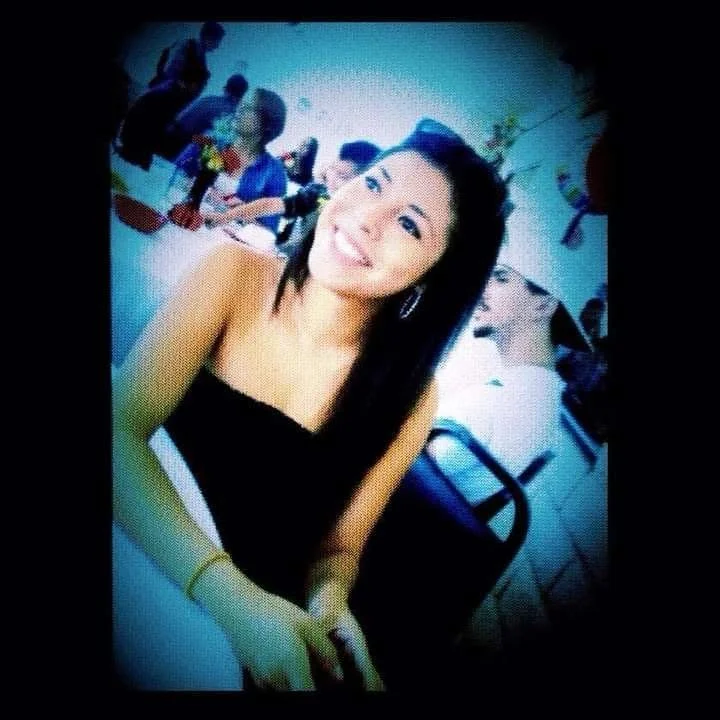 A young woman smiling at a party, sitting at a table with other people in the background.