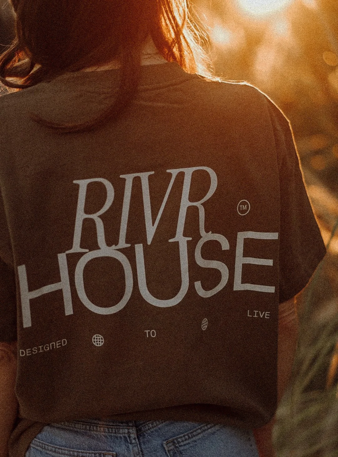 A person wearing a black T-shirt with the words 'RVR HOUSE' in large white letters on the back, standing outdoors during sunset.