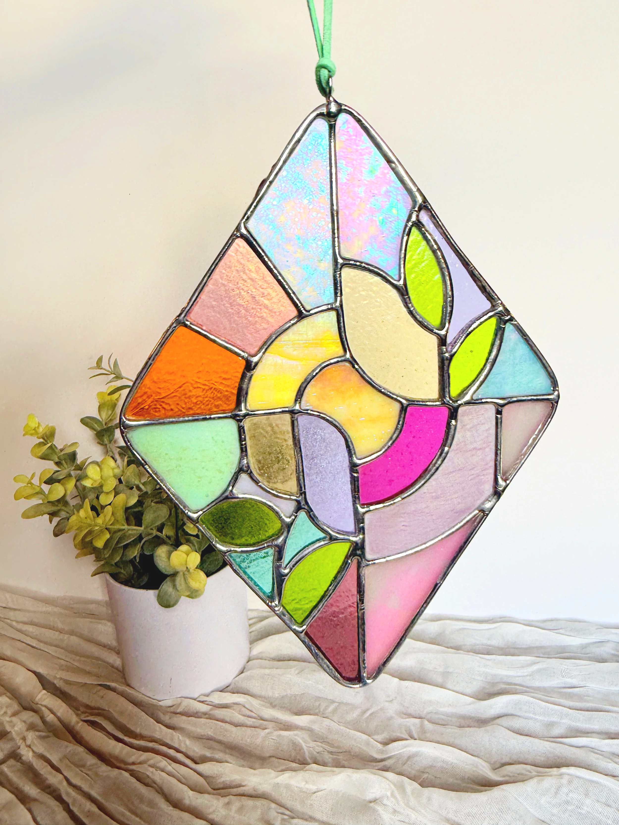 Stained glass ornament shaped like a Monstera leaf hanging outdoors with greenery background.