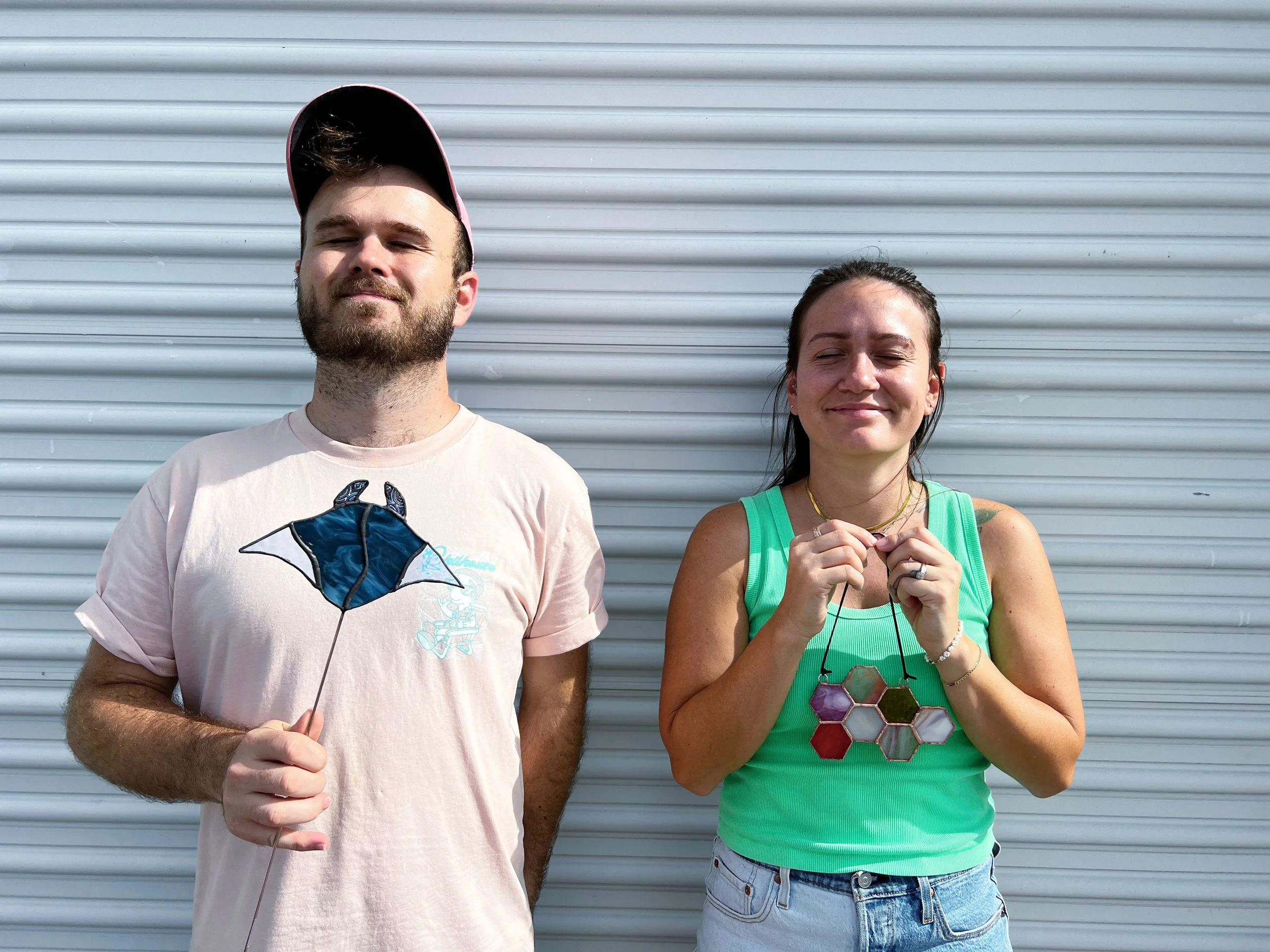 Two people standing in front of a corrugated metal wall, each holding a stained glass ornament. The man on the left wears a light pink shirt and pink baseball cap, holding a manta ray ornament. The woman on the right wears a green tank top and jeans, holding a hexagonal pattern ornament.