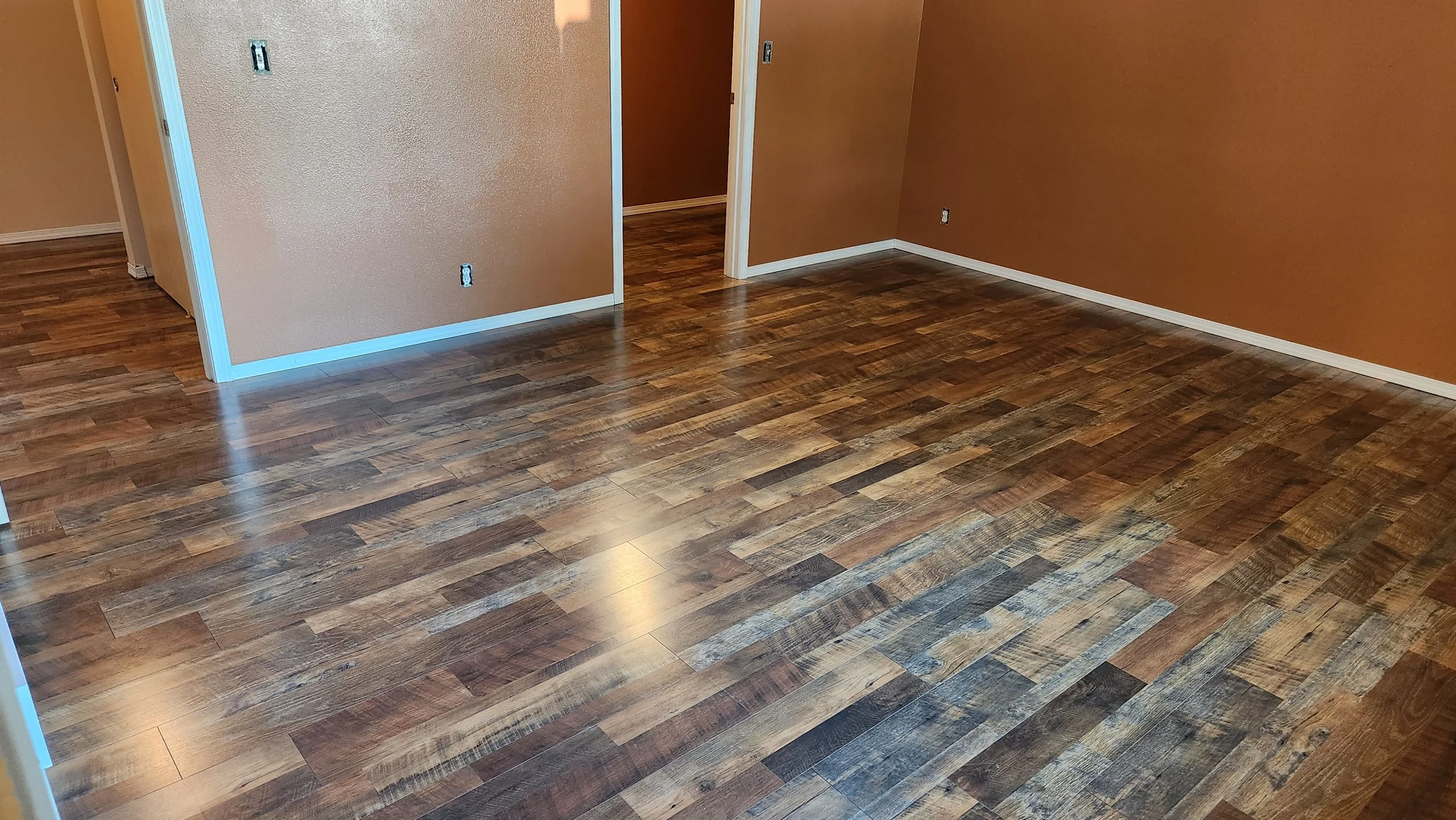 An empty room with hardwood flooring and beige walls, some of which have electrical outlets, with an open doorway leading to another room.