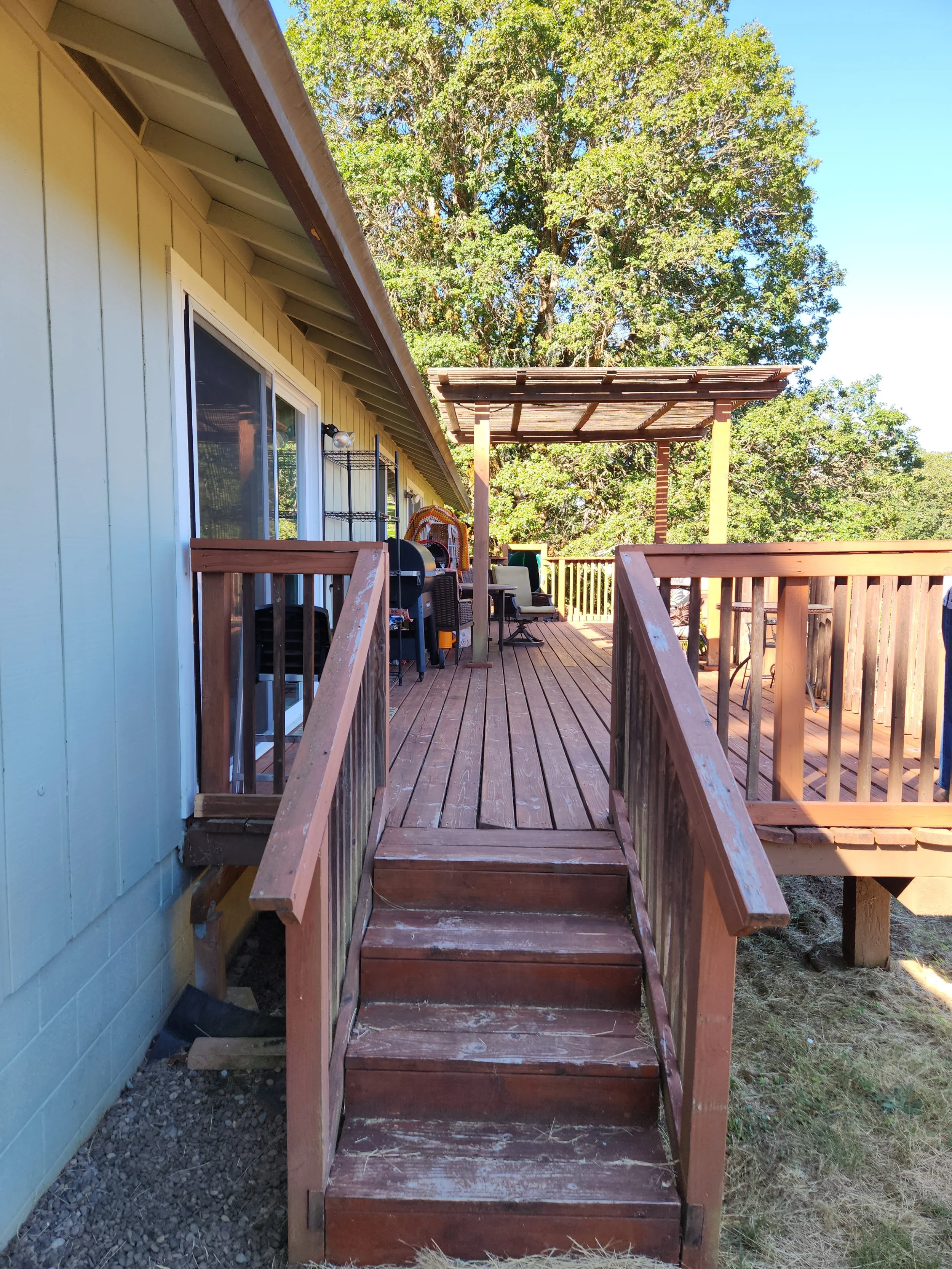 Wooden deck detached to house with stairs and outdoor furniture, with a large tree and a clear blue sky in the background. Before picture of a rotted deck