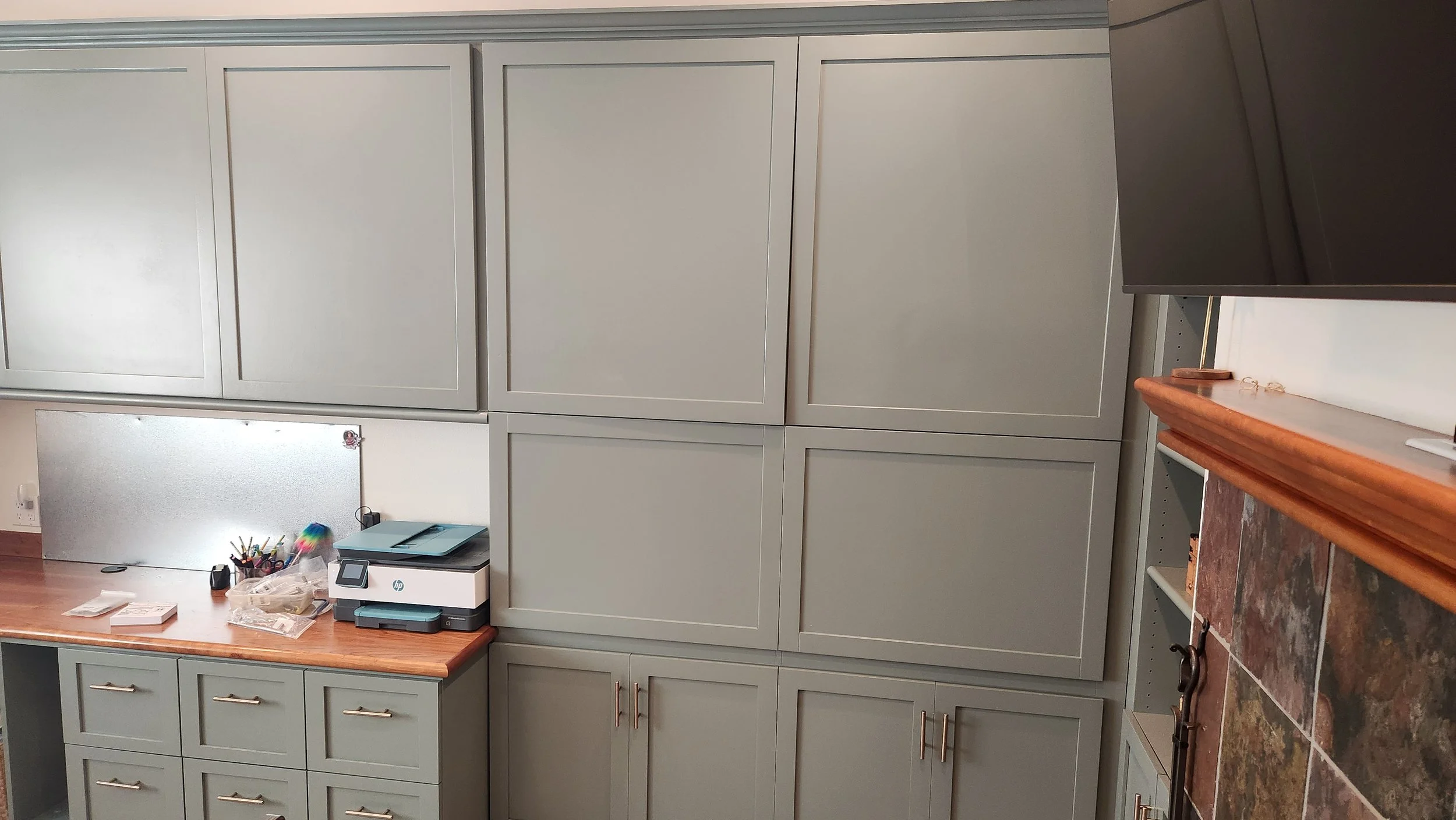 Office with green/gray cabinets, a wooden desktop, a printer and office supplies on the counter, a stainless steel panel, and a tiled surface.