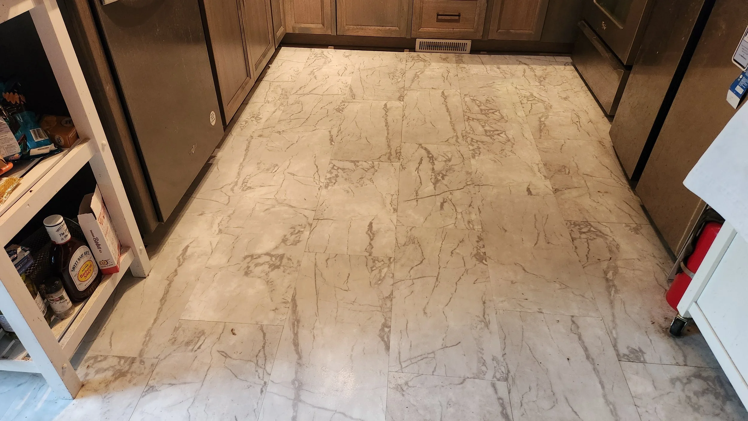 installed the flooring.  Surrounding cabinets and appliances are visible, including a stainless steel refrigerator on the left, a stove on the right, and a white shelving unit with various items on the far left.