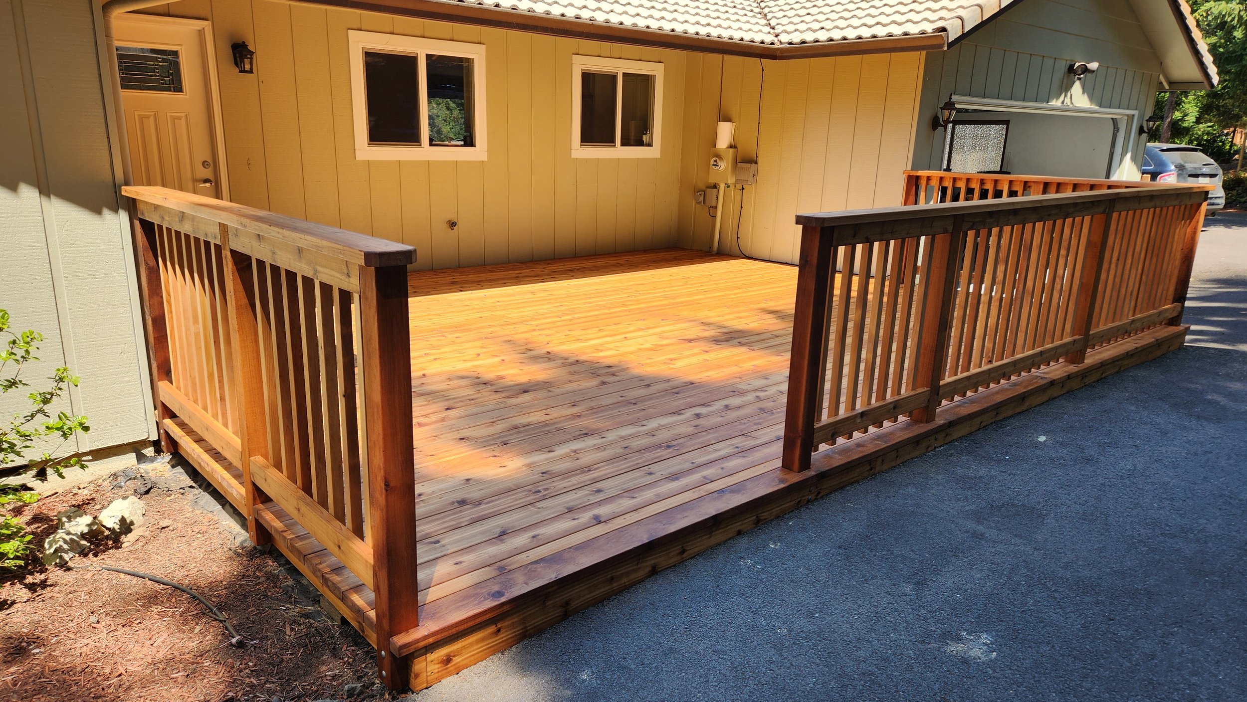 Newly built western red cedar deck with railing attached to the front of a yellow house with two windows and a door, with a driveway and parked cars in the background.