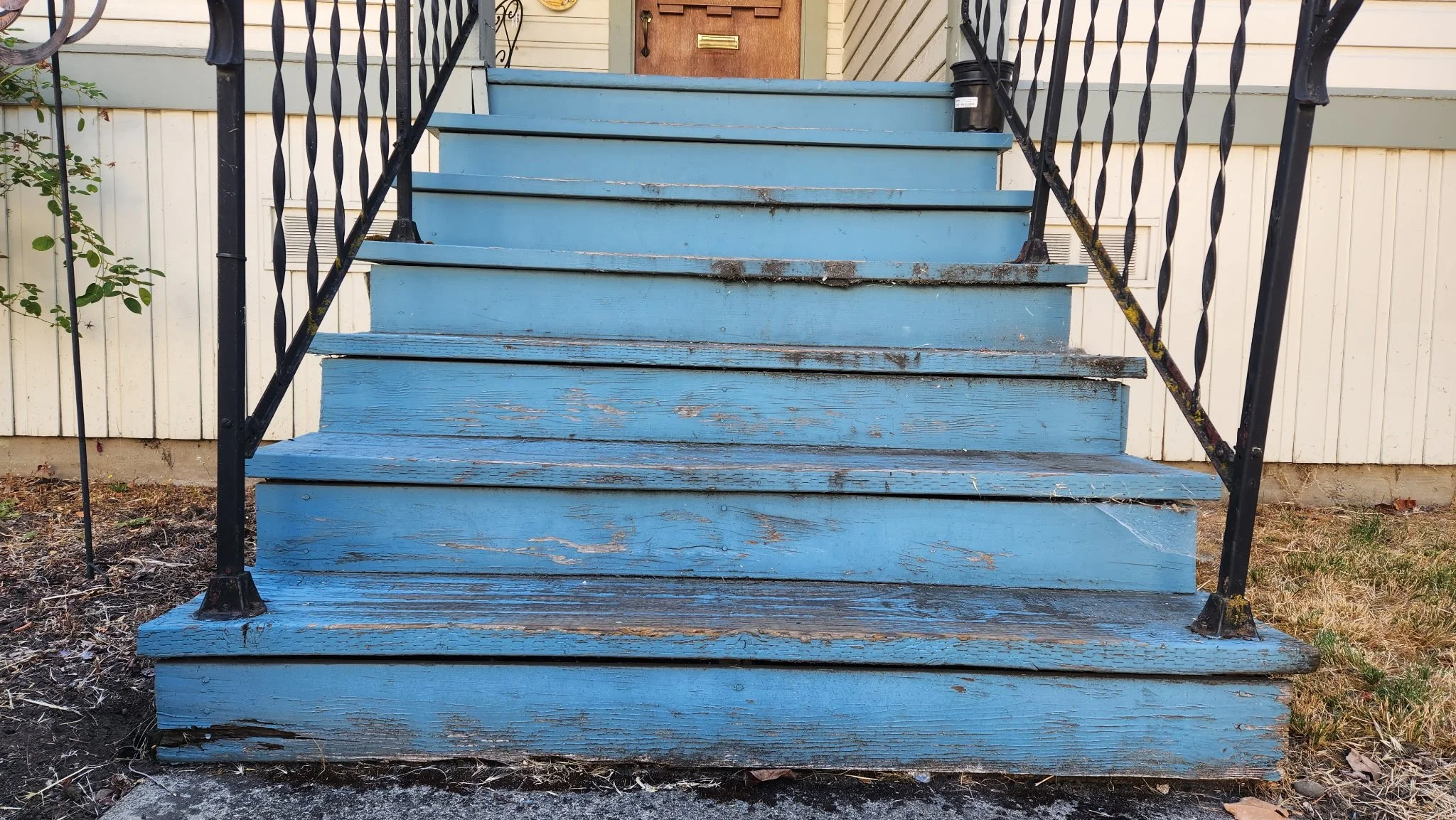 Front view of a worn blue wooden staircase with black metal railings leading to a front door of a house. The staircase shows signs of peeling paint and weathering.