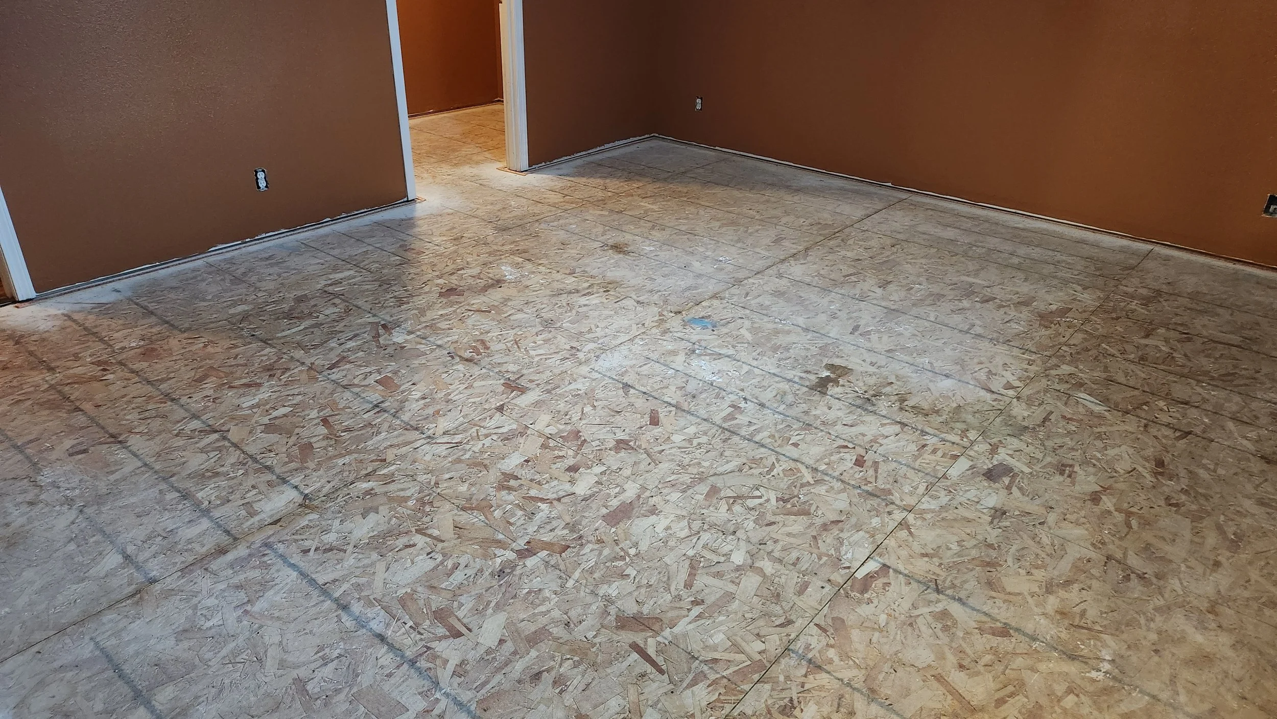 Interior room with unfinished floor and brown walls, doorway in the background, multiple electrical outlets on walls. Carpet was pulled to prep for the installation of hardwood flooring.