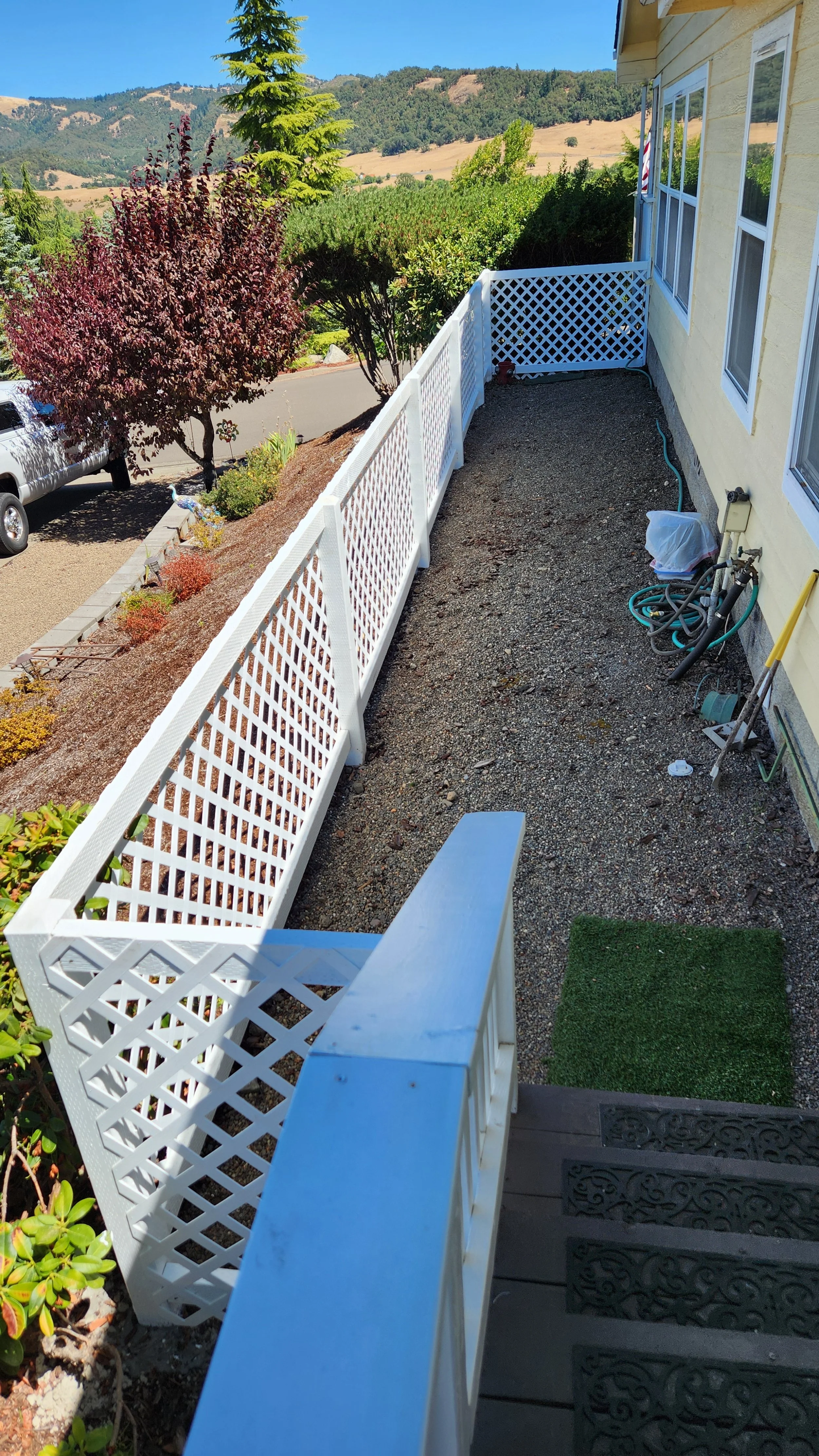 Finished dog run white lattice railing, steps with black patterned mats, and a yard with shrubs and trees on a sunny day with hills in the background.