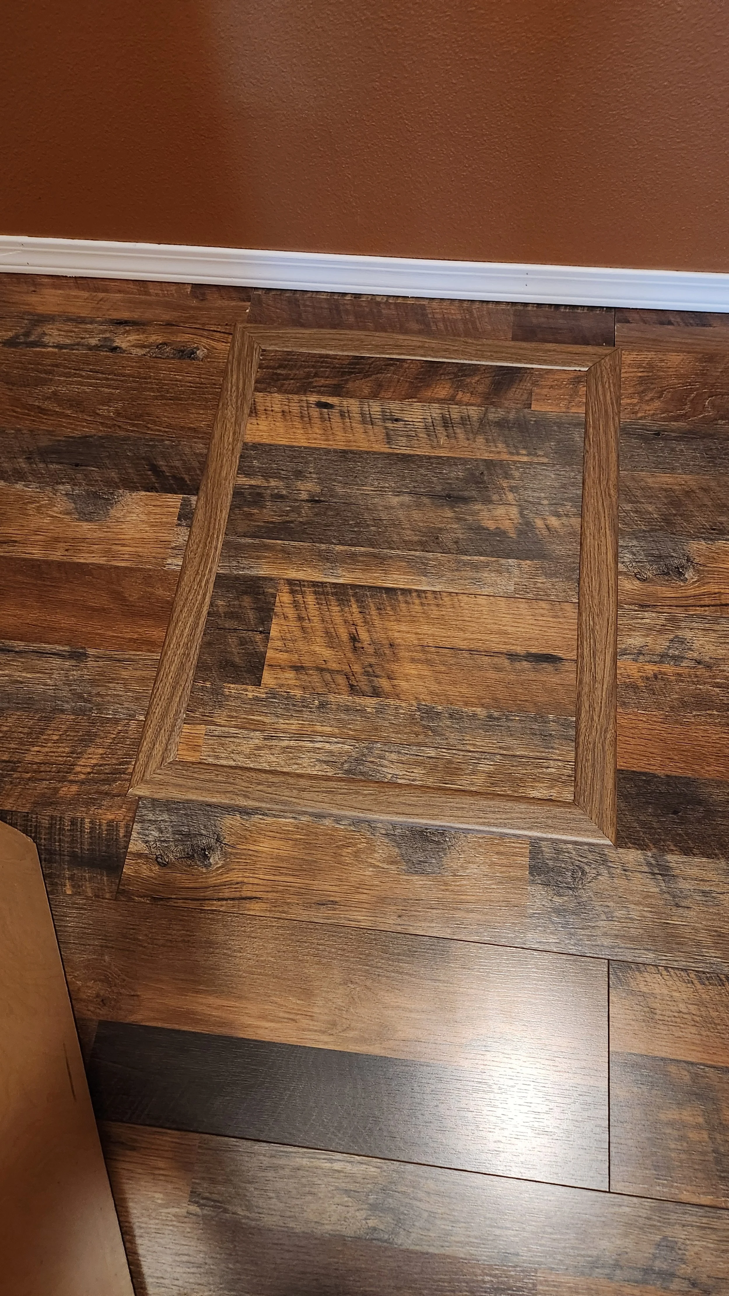 Wooden floor with a matching crawlspace access panel and a wall with a white baseboard in the background.