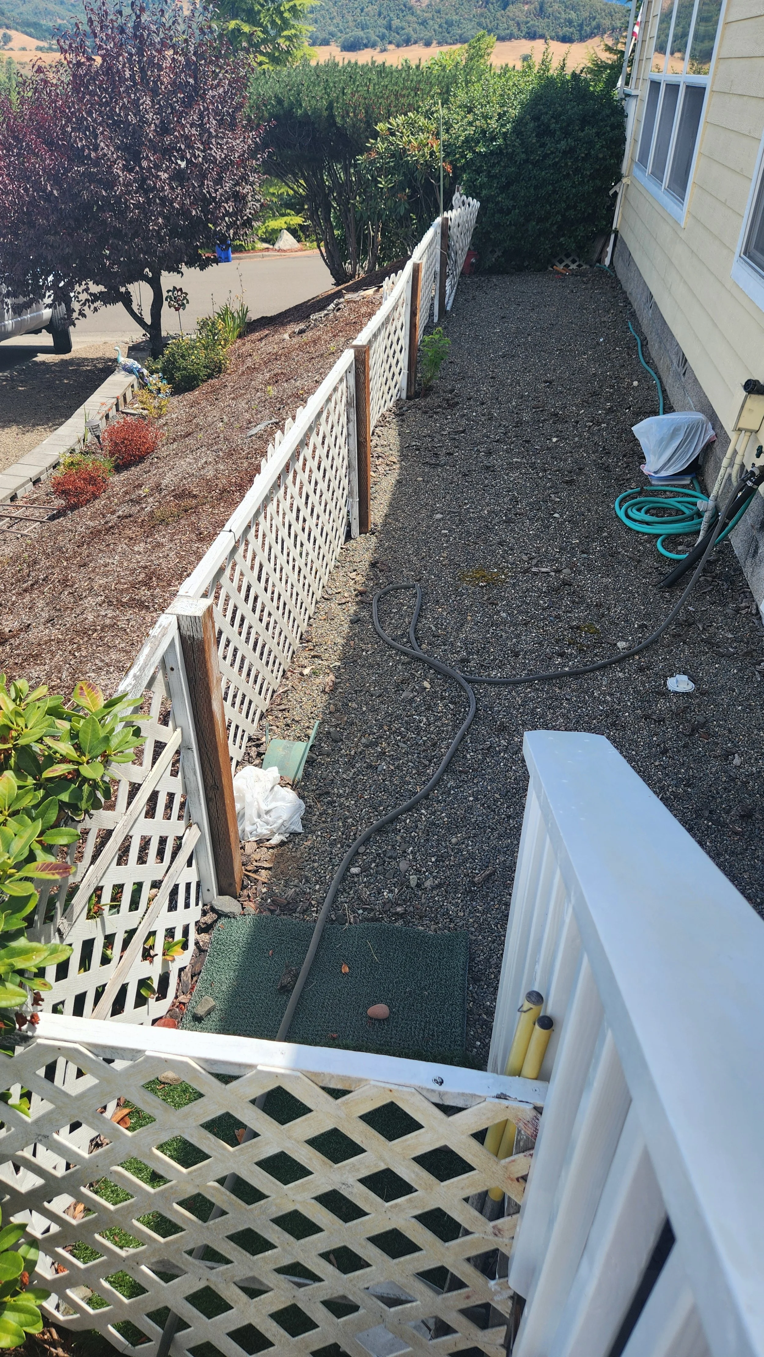 A backyard side yard that needs to be rebuilt with white lattice fencing, gravel ground, and garden hoses, next to a yellow house and overlooking green hills.